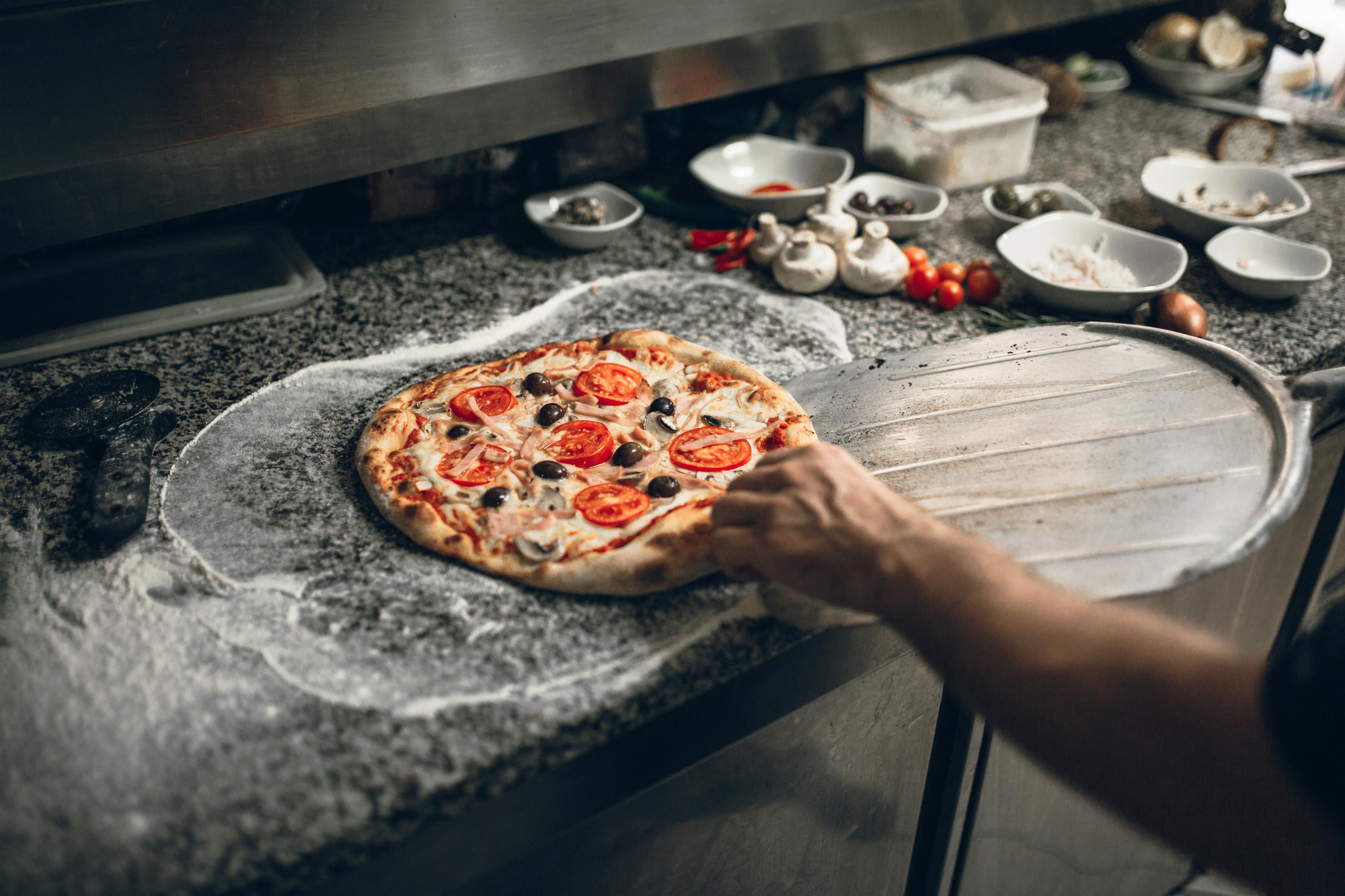 Eine frisch gebackene Pizza mit Tomaten und Oliven wird von der Theke genommen. Eine Hand hält den Pizzaschieber.