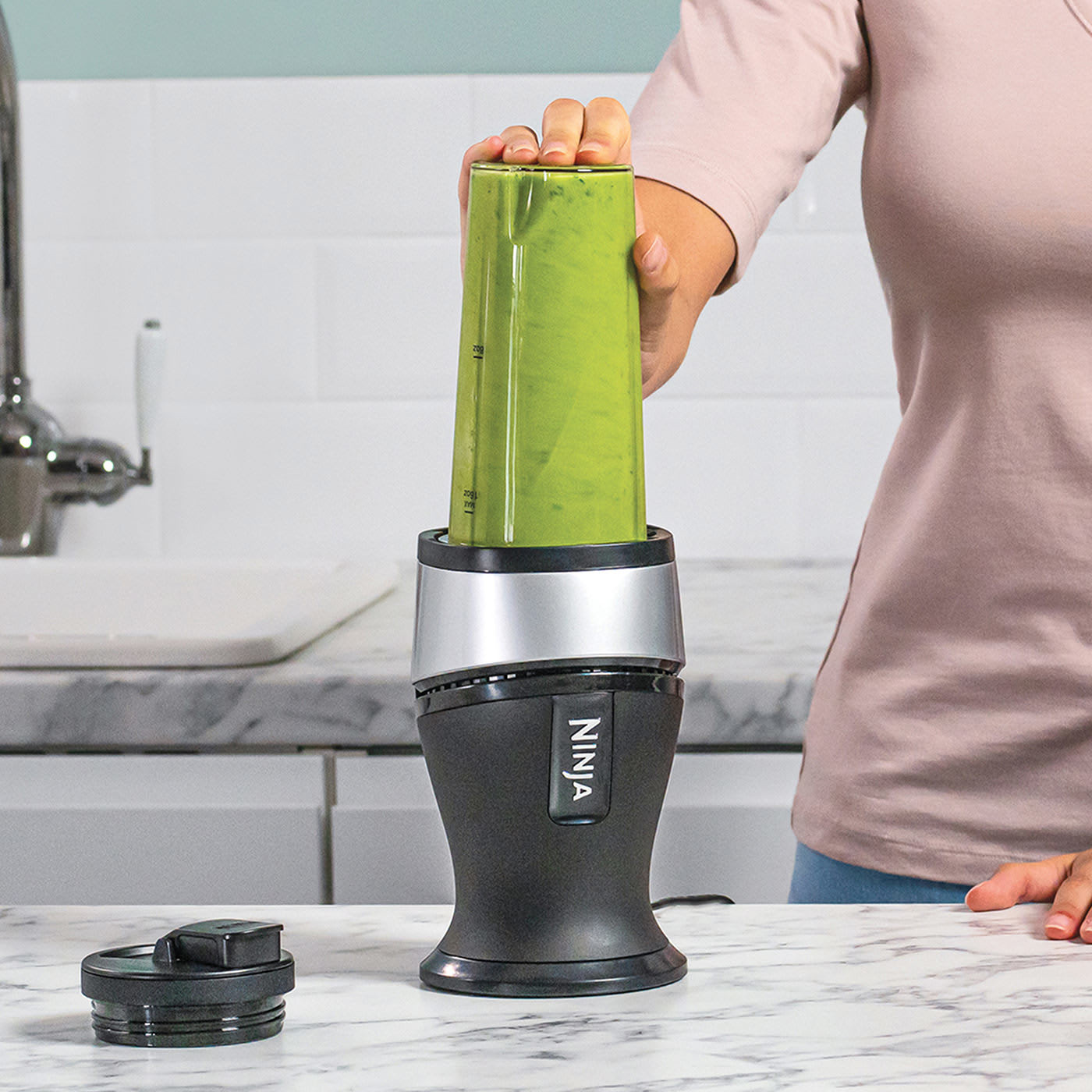Une femme tient un récipient de smoothie vert sur un blender Ninja. Le blender est noir et argenté.