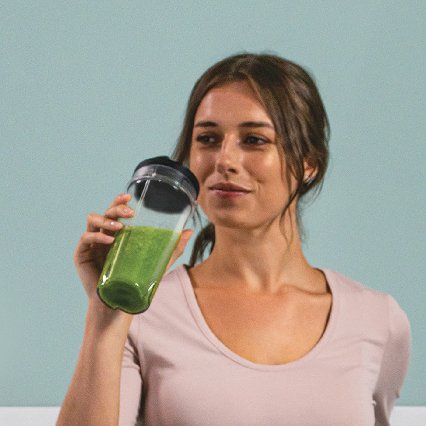 Une femme tient un smoothie vert dans un gobelet en plastique transparent. Elle sourit et a les cheveux bruns.