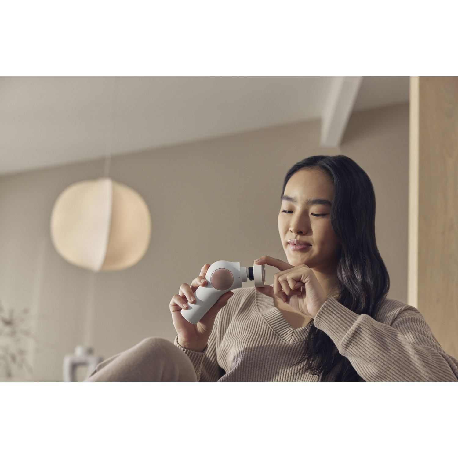 Mujer sosteniendo un dispositivo de belleza blanco y rosa, interactuando con él. Fondo neutro, tonos cálidos.
