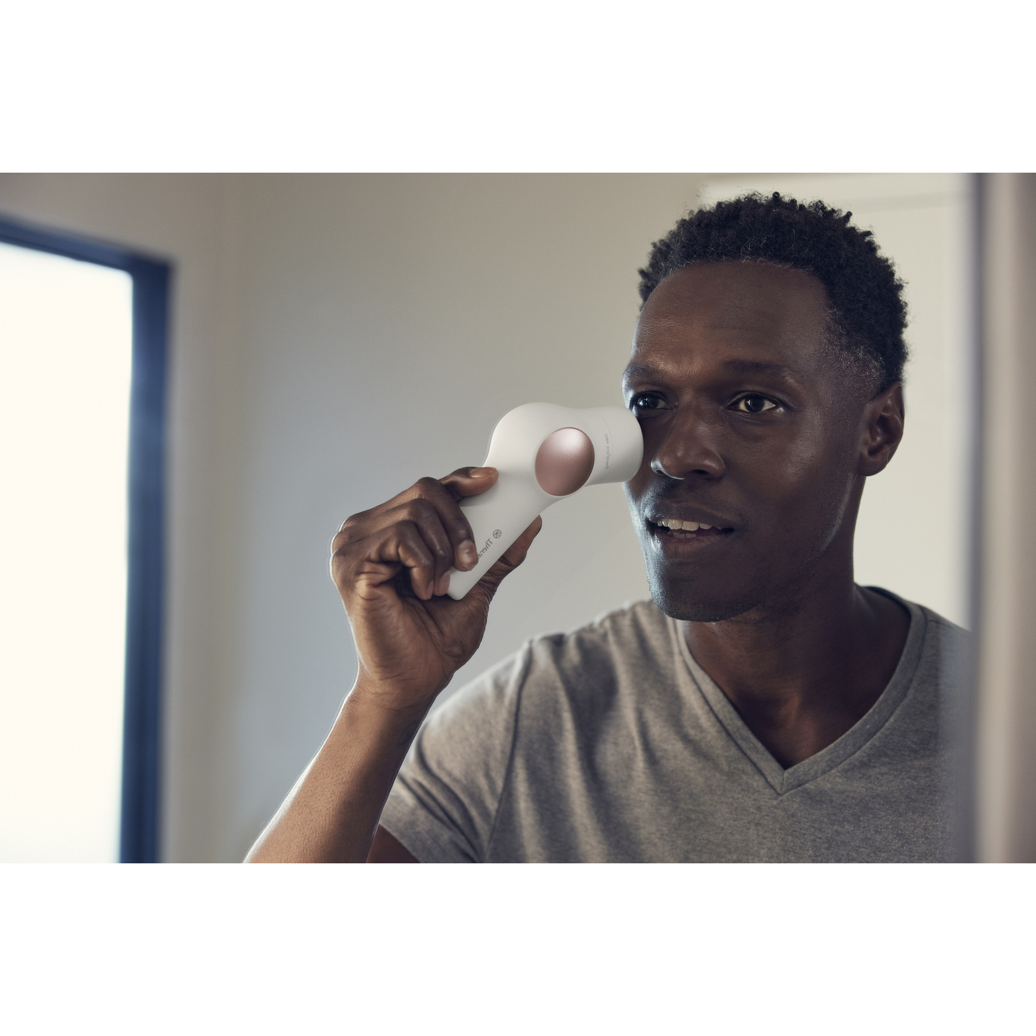Hombre sostiene dispositivo limpieza facial blanco en ojo, mirándose espejo.