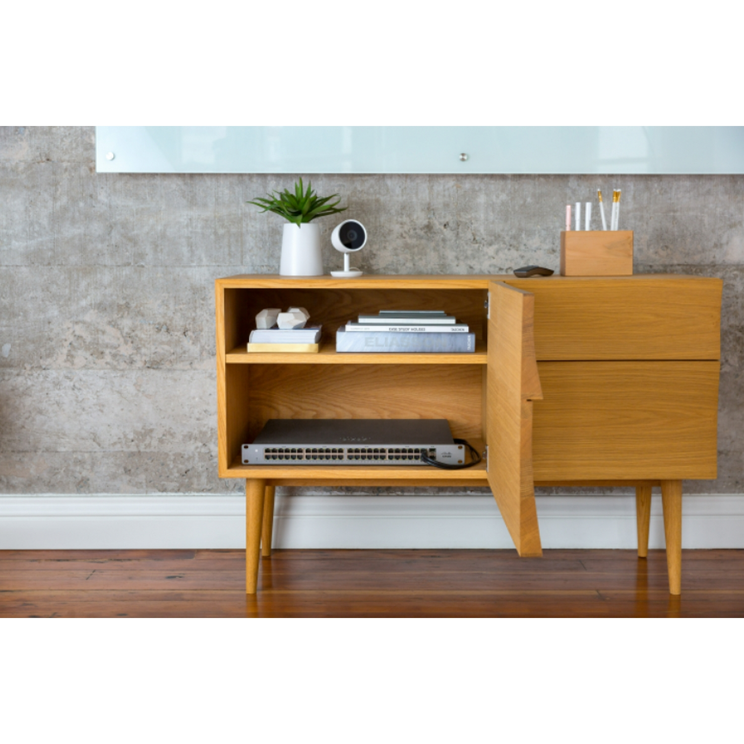 Un mueble de madera con planta, cámara, libros y un switch de red.