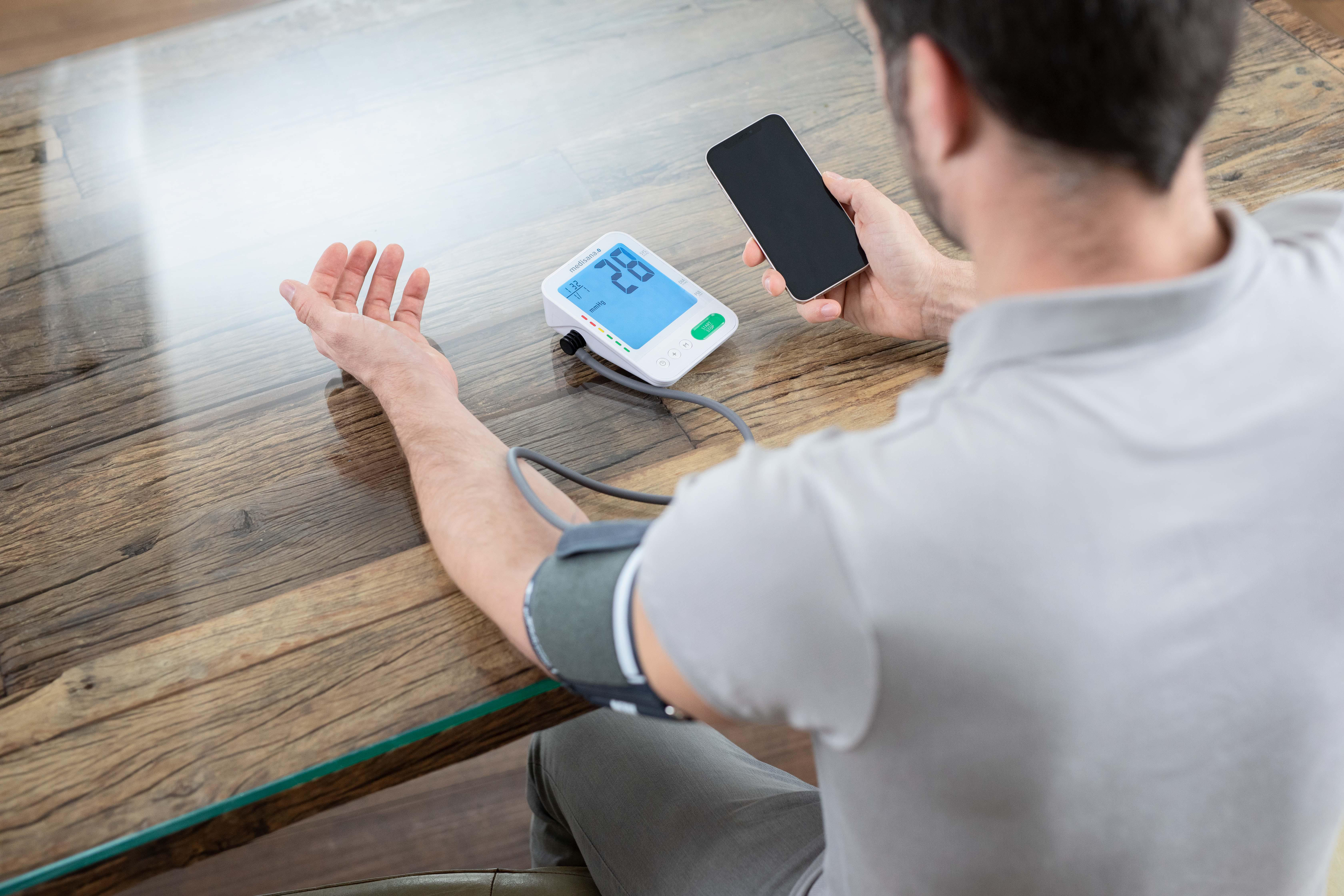 Person misst Blutdruck mit Gerät und hält Smartphone.