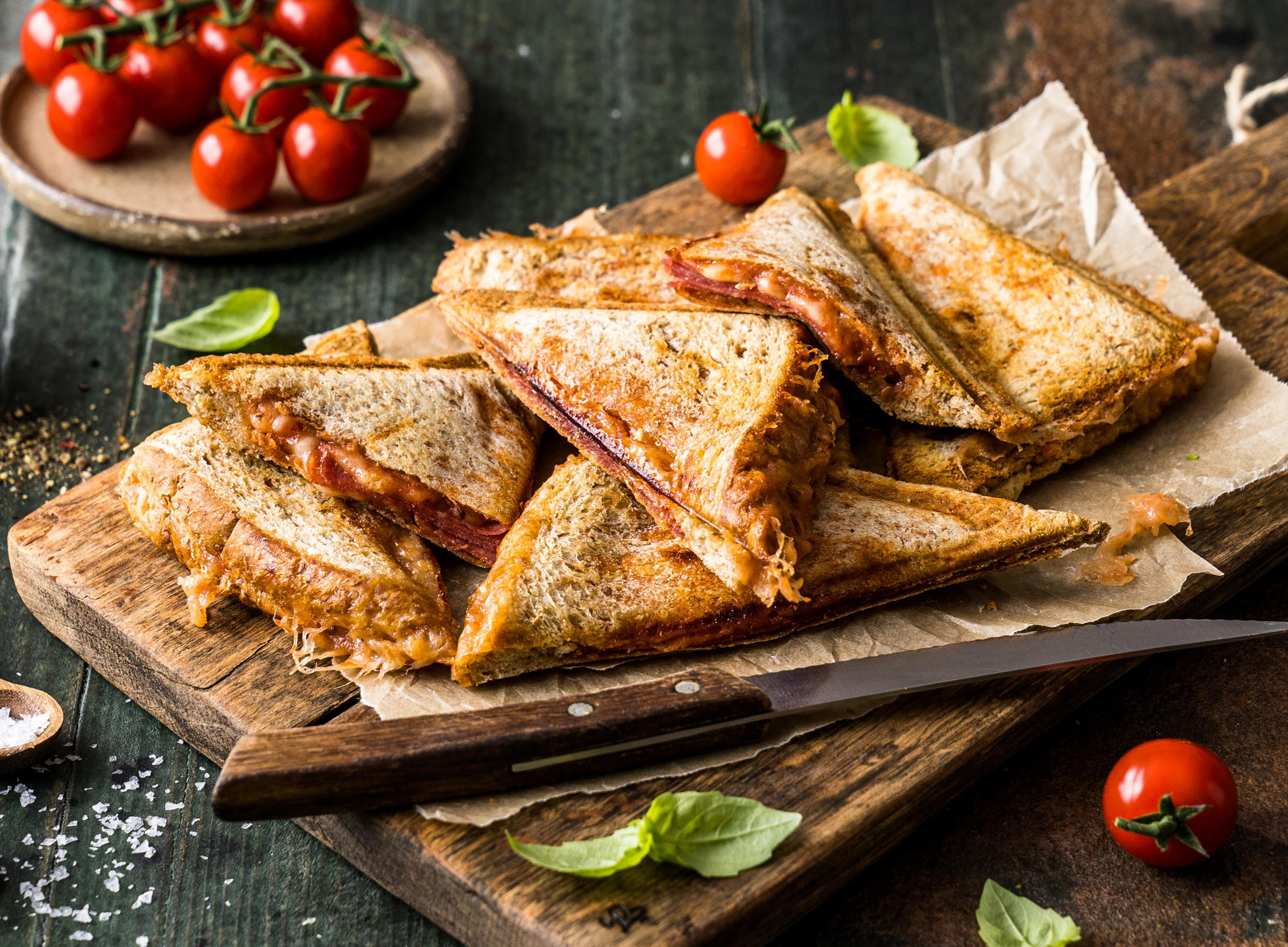 Gegrillte Käsesandwiches auf einem Holzbrett, mit Tomaten und einem Messer.