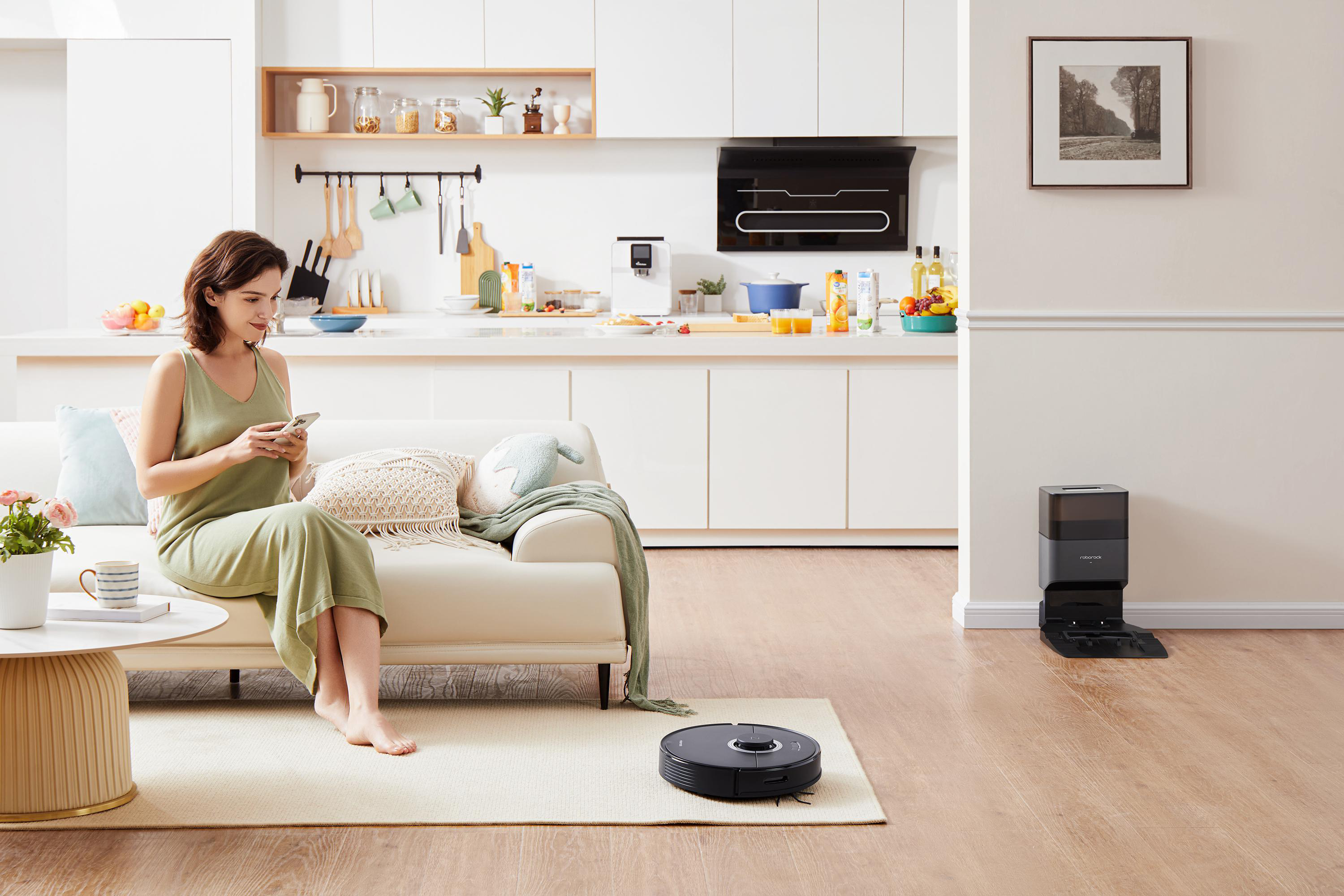 Frau auf Couch mit Handy, Roboterstaubsauger auf Teppich, Küche im Hintergrund.
