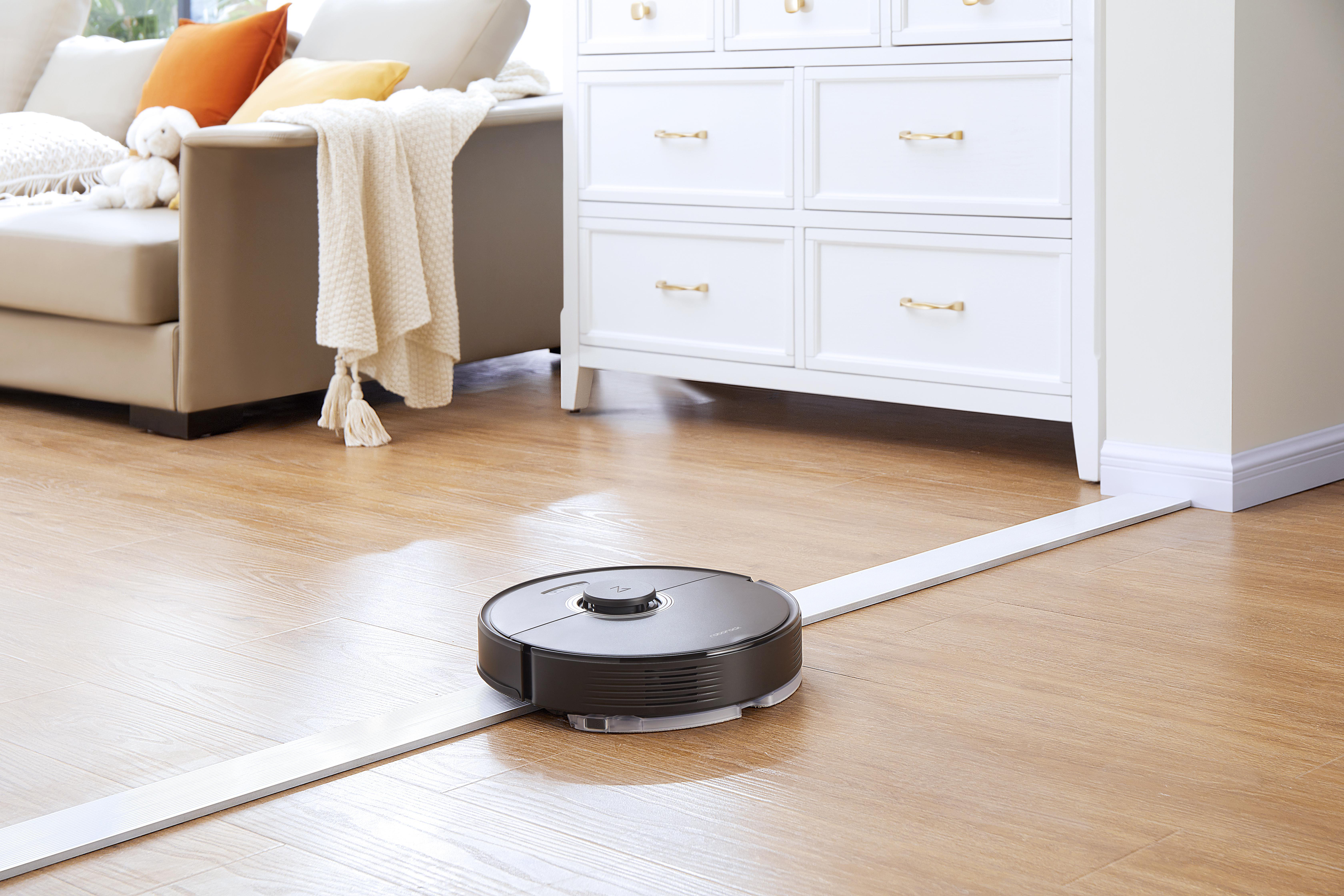 Ein schwarz-silberner Roboter-Staubsauger reinigt einen Holzboden mit Möbeln im Hintergrund.