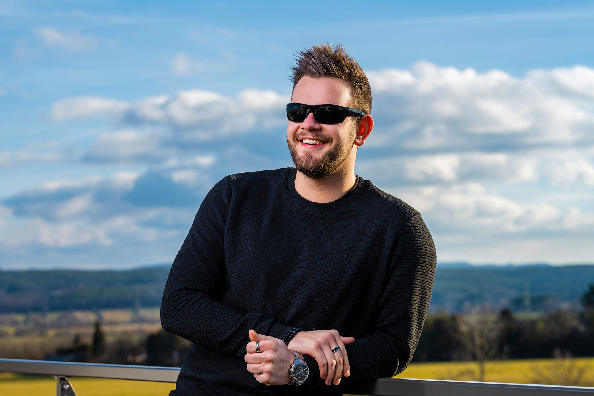 Ein lächelnder Mann mit Sonnenbrille und schwarzem Pullover, der sich im Freien an ein Metallgeländer lehnt.