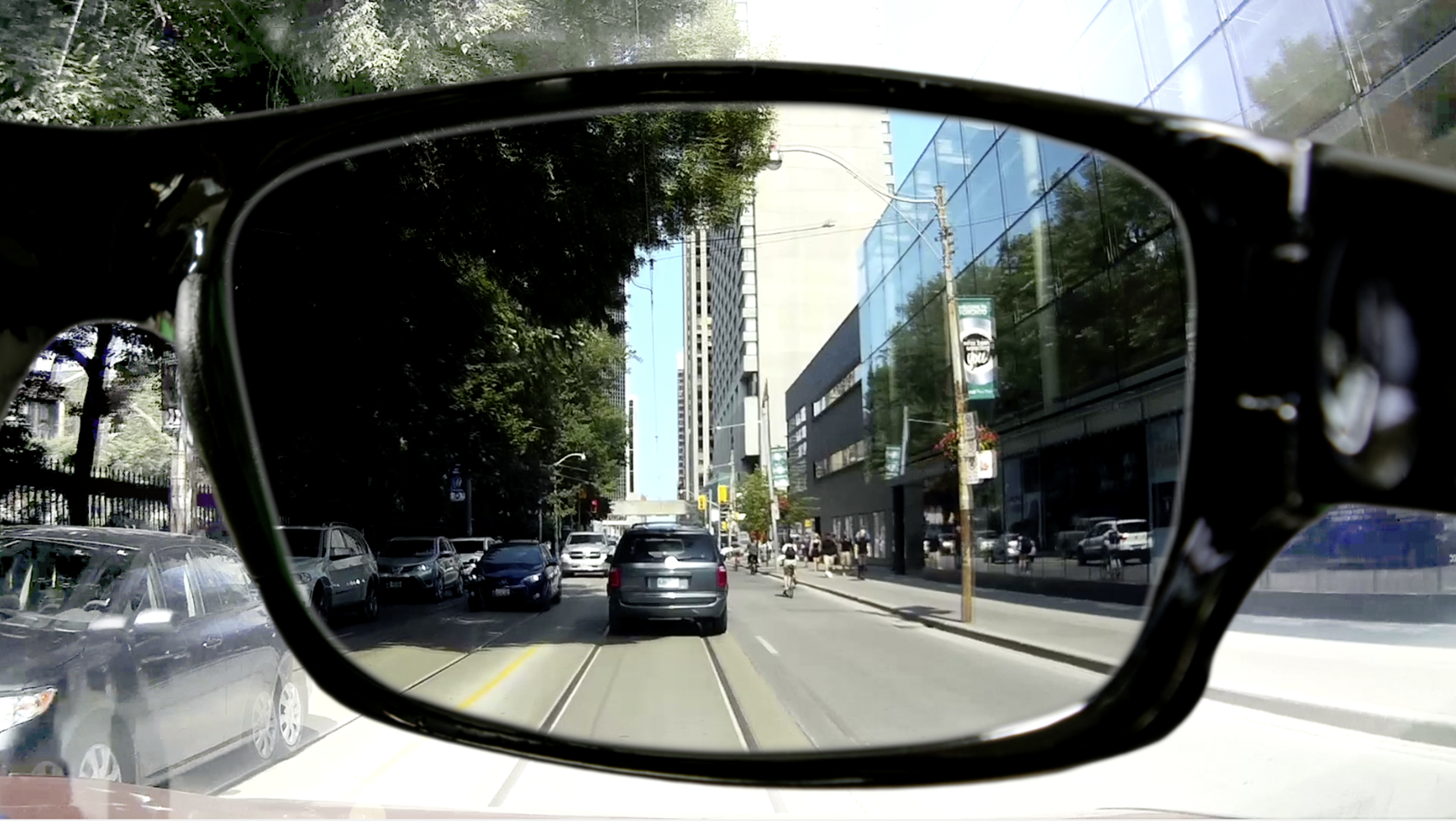 Blick auf eine Stadtstraße durch eine schwarz gerahmte Brille. Autos und Gebäude sind sichtbar.