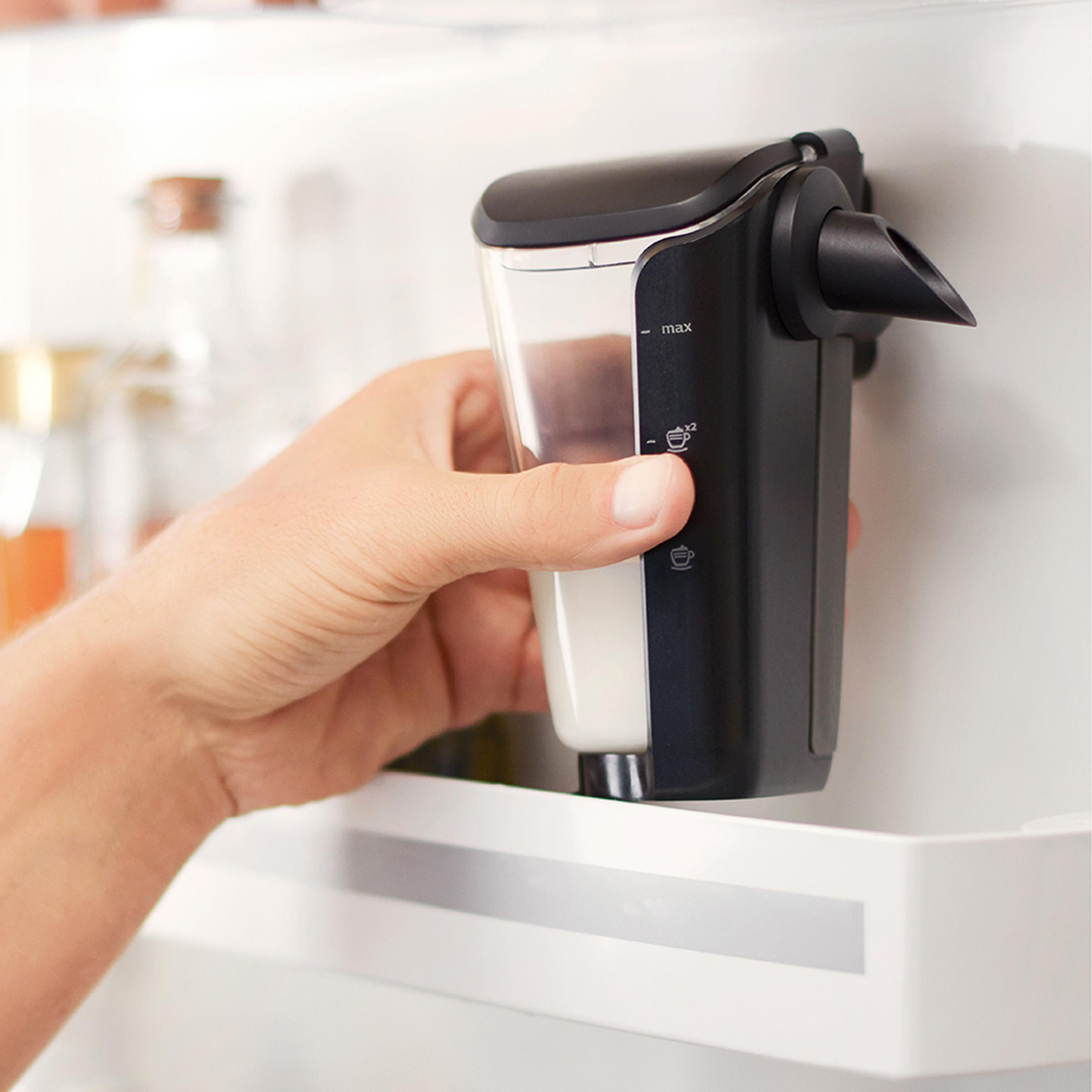 Hand hält einen schwarzen Milchaufschäumer, befestigt an einem weißen Kühlschrank, mit Milch darin.