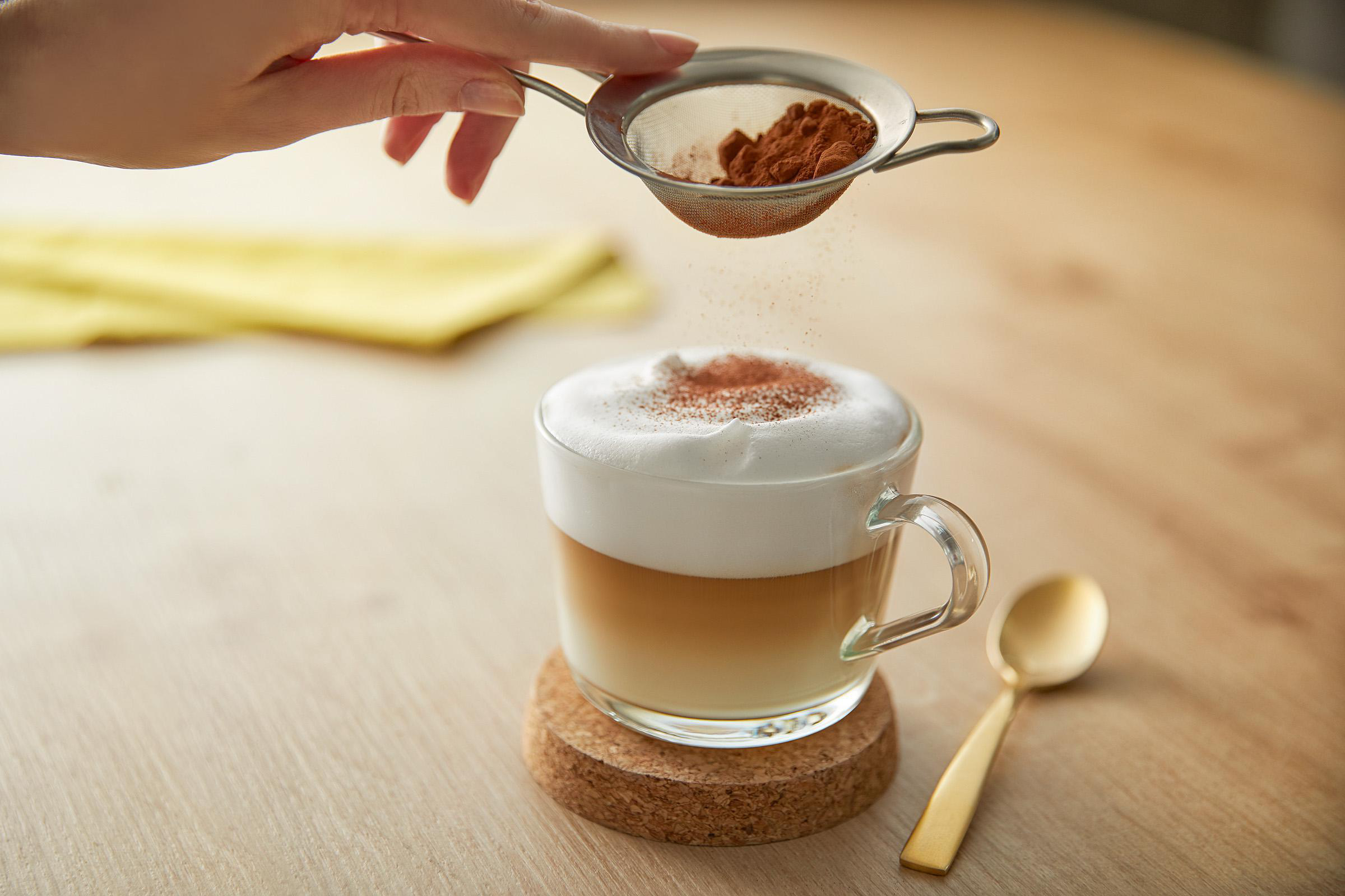 Eine Hand streut Kakaopulver auf einen Latte mit Schaum. Ein Löffel liegt neben der Tasse auf einem Korkuntersetzer.
