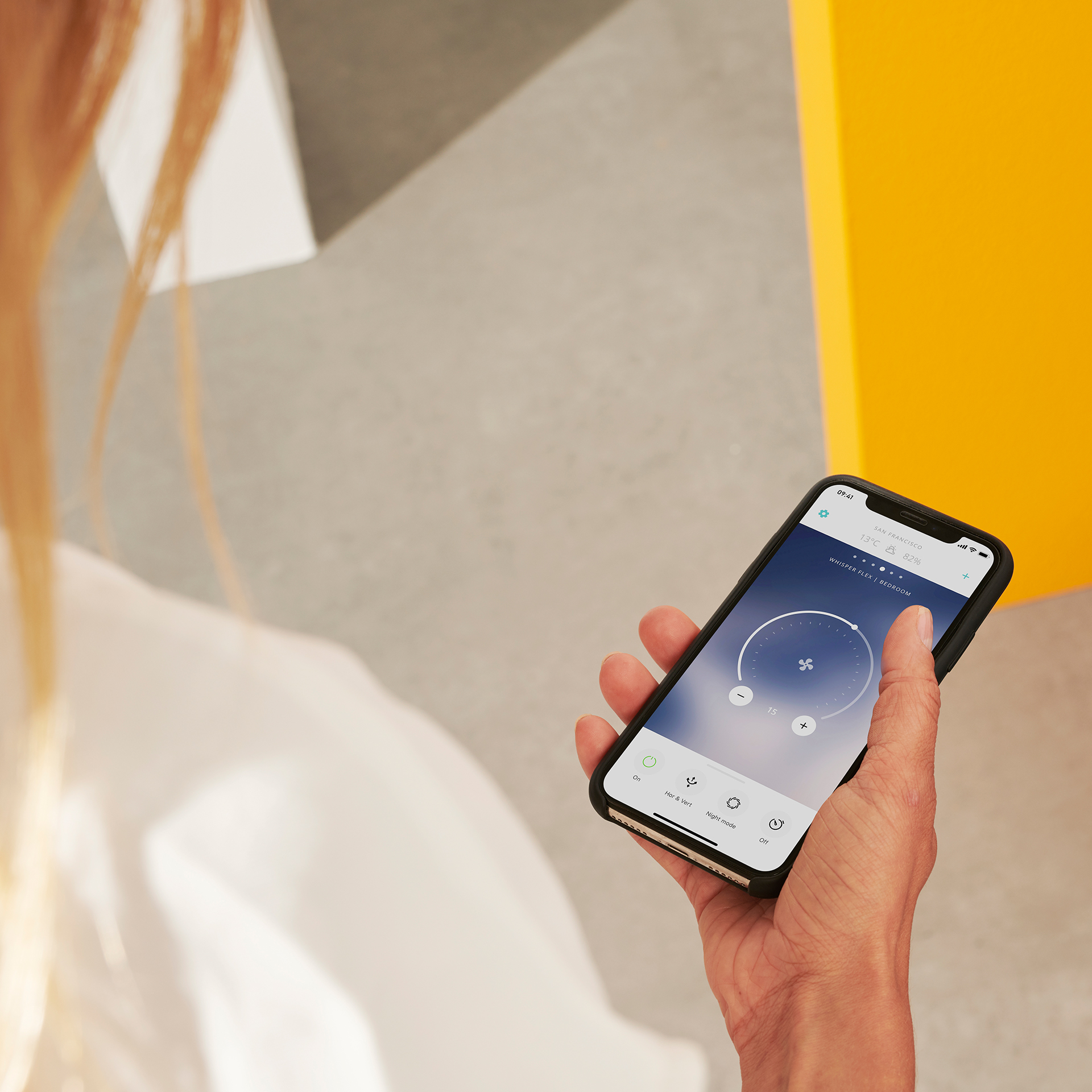 Een persoon die een telefoon vasthoudt met een ventilatorbedieningsinterface, in de buurt van een gele muur.