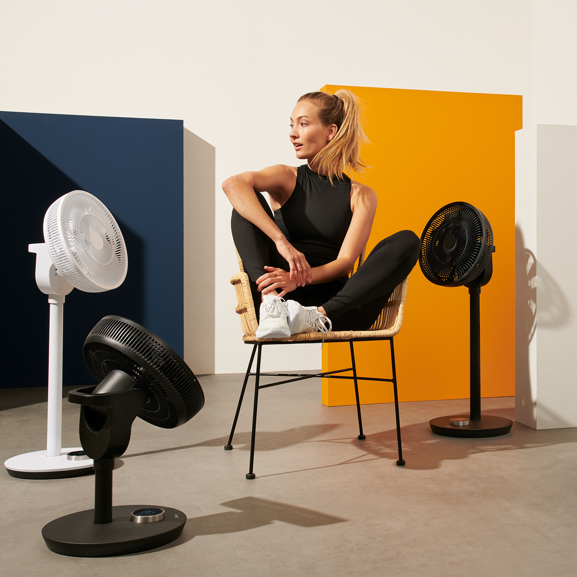 A woman sits in a chair surrounded by fans. Two black and one white fan are placed in the room.