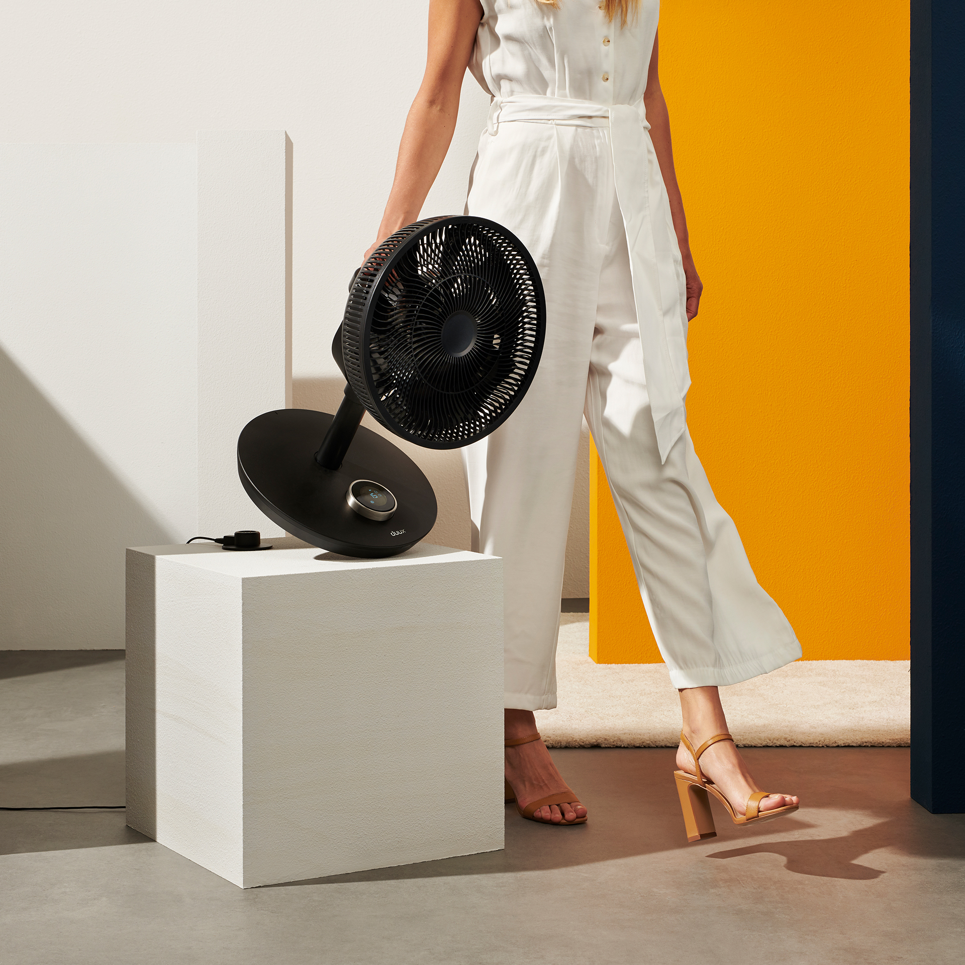 Woman holding a black fan on a white cube, with yellow and blue background.