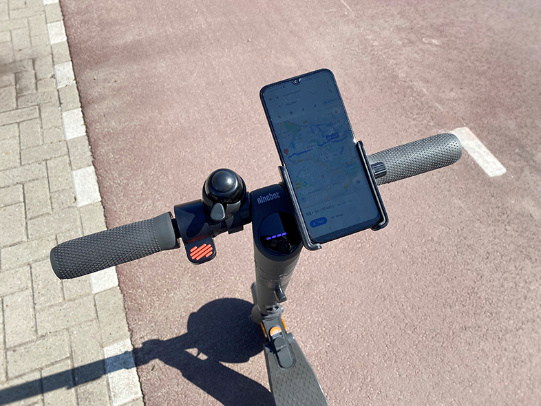 Robogó kormánya telefontartóval. A telefonon egy térkép látható. Járda és út látható.