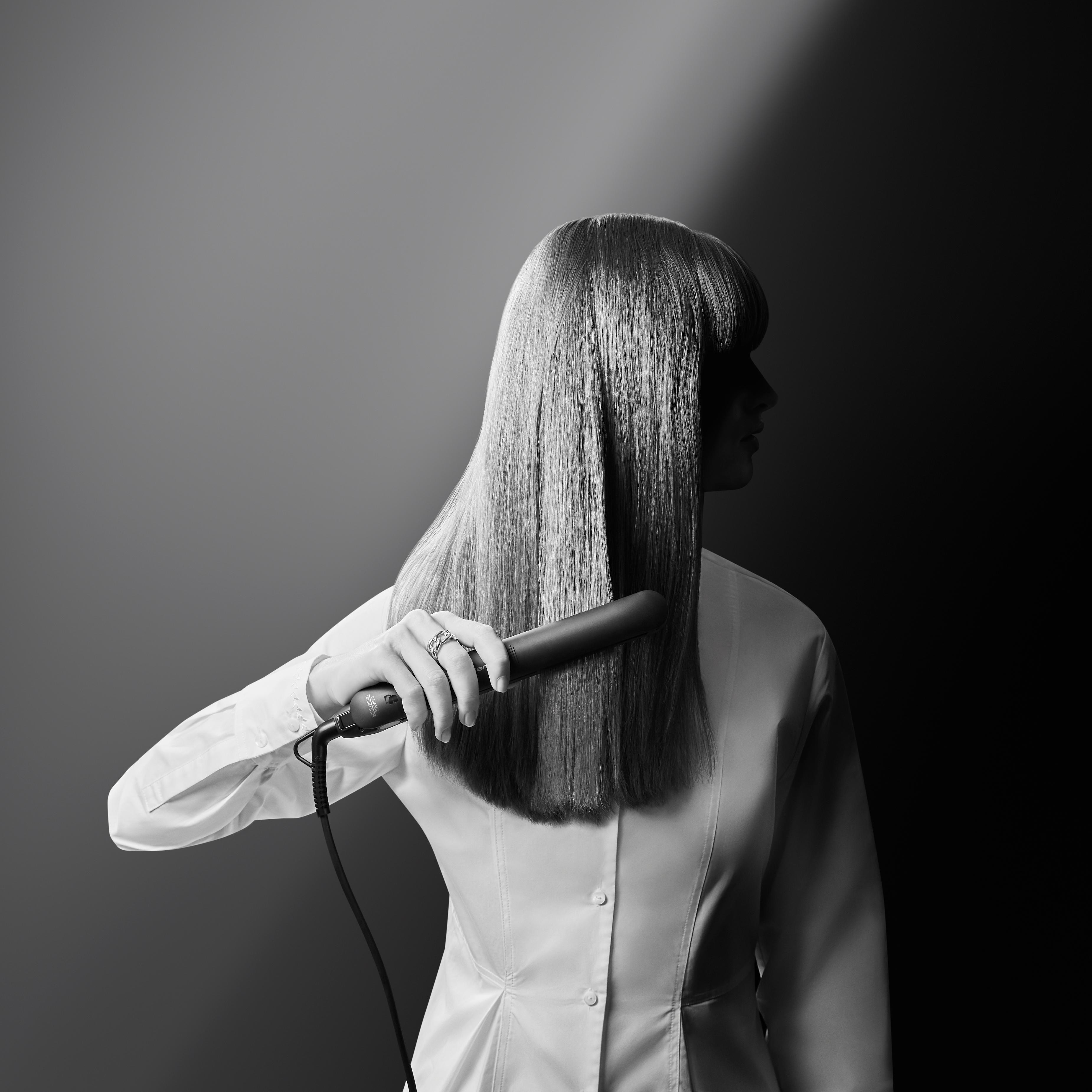 Eine Frau mit langen, glatten blonden Haaren hält ein Glätteisen. Der Hintergrund ist dunkel.