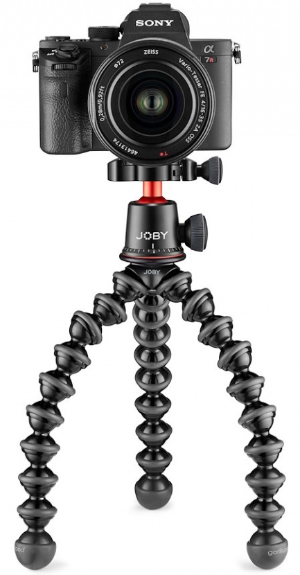 Een zwarte camera met een lens staat bovenop een flexibel statief. Het statief heeft een rode kogelkop en gebogen poten.
