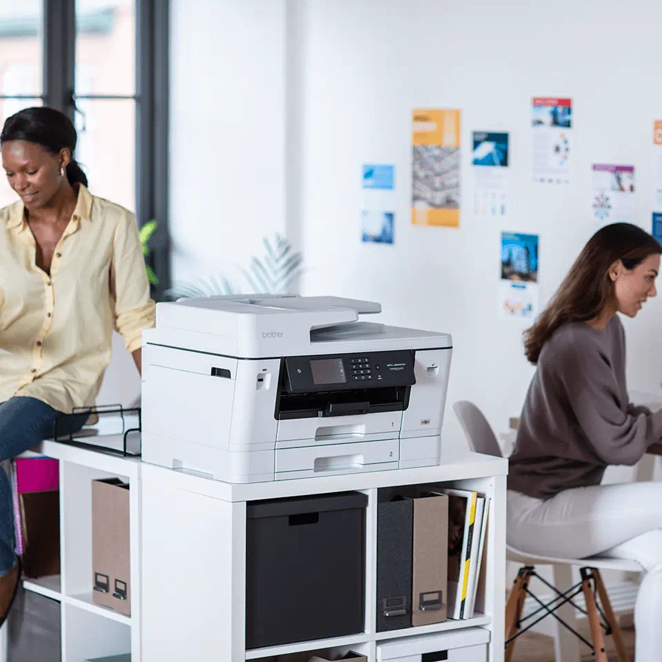 Due donne in un ufficio. Una stampante è su uno scaffale. Fogli sulla parete.
