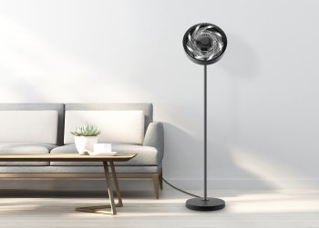 Schwarzer Ventilator in einem Wohnzimmer. Sofa, Tisch mit Pflanze und weiße Wand sichtbar.