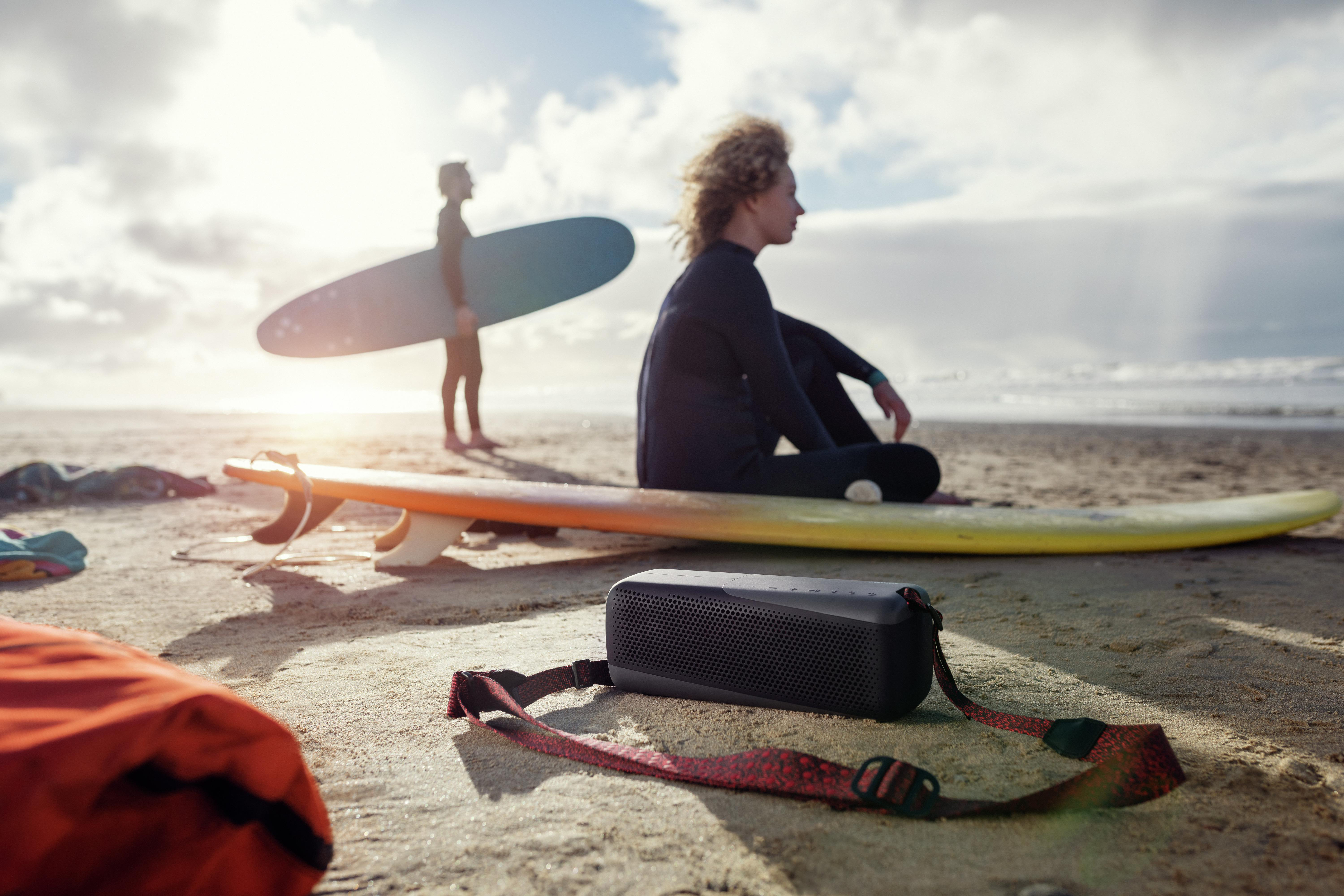 Strandszene mit zwei Surfern und einem Lautsprecher. Ein Surfer steht, einer sitzt auf einem Brett.