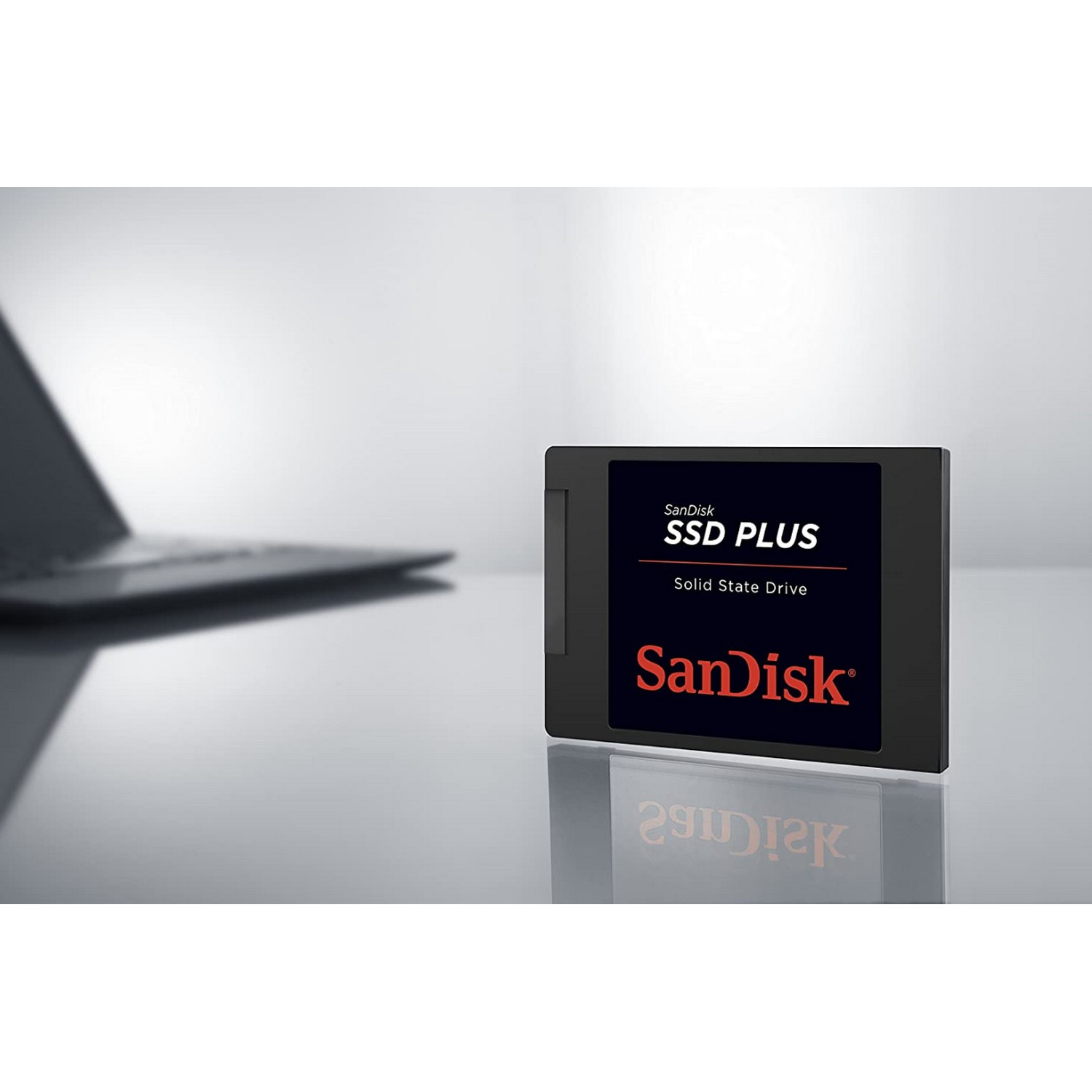 Un SSD SanDisk PLUS negro y plateado. El SSD está sobre una superficie reflectante.