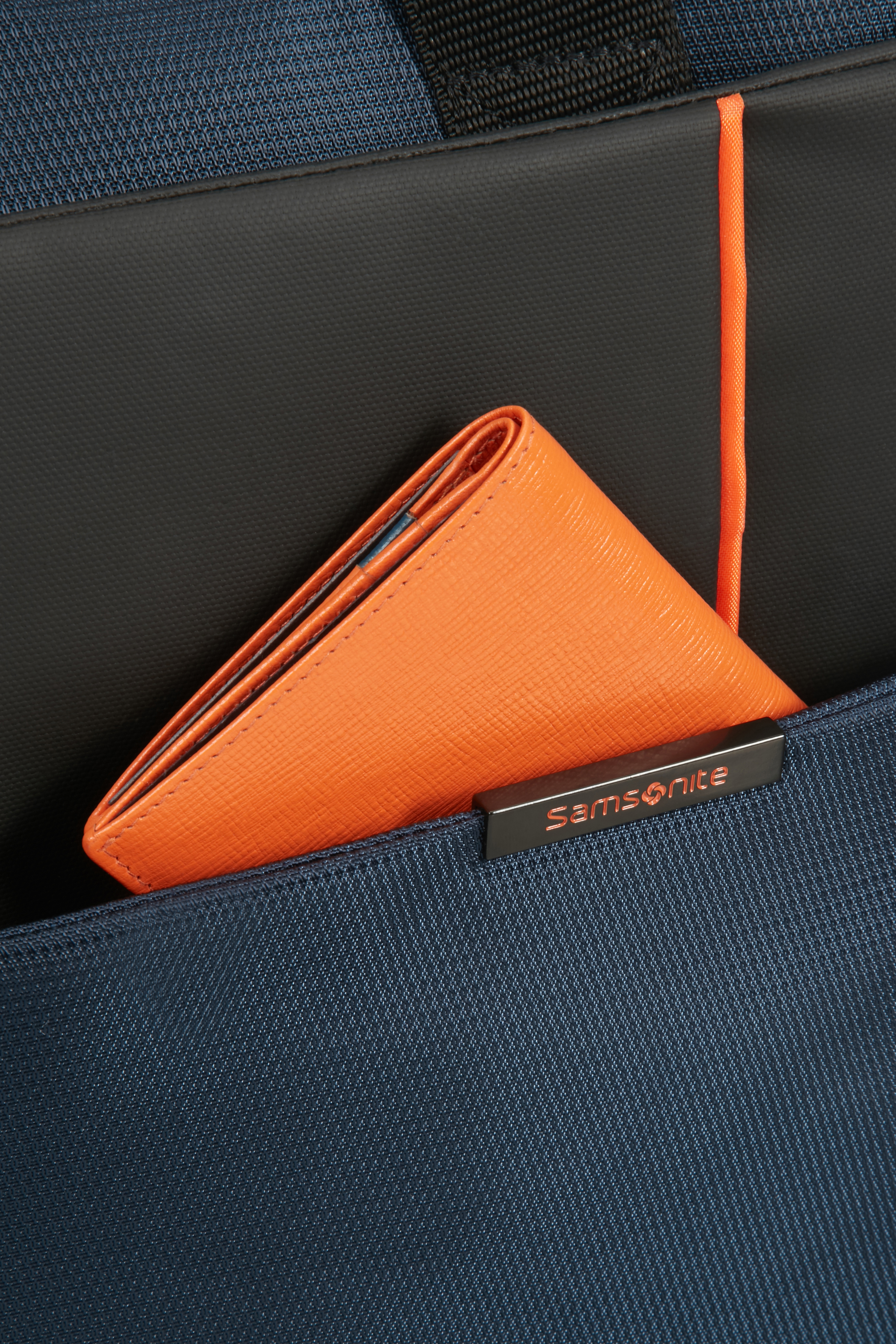 Mavi bir Samsonite çanta cebinde turuncu bir cüzdan. Gümüş bir Samsonite etiketi görünür.