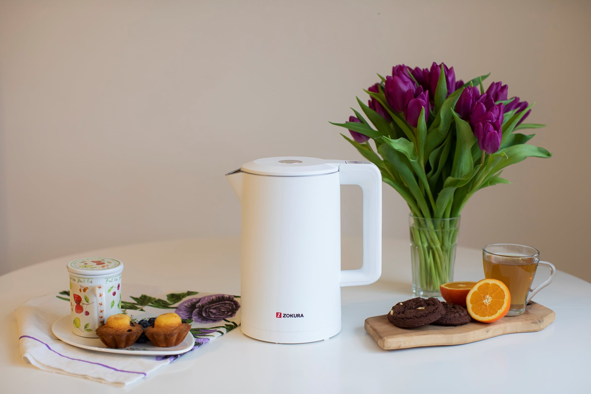 Fehér elektromos vízforraló lila tulipánokkal és muffinokkal az asztalon.