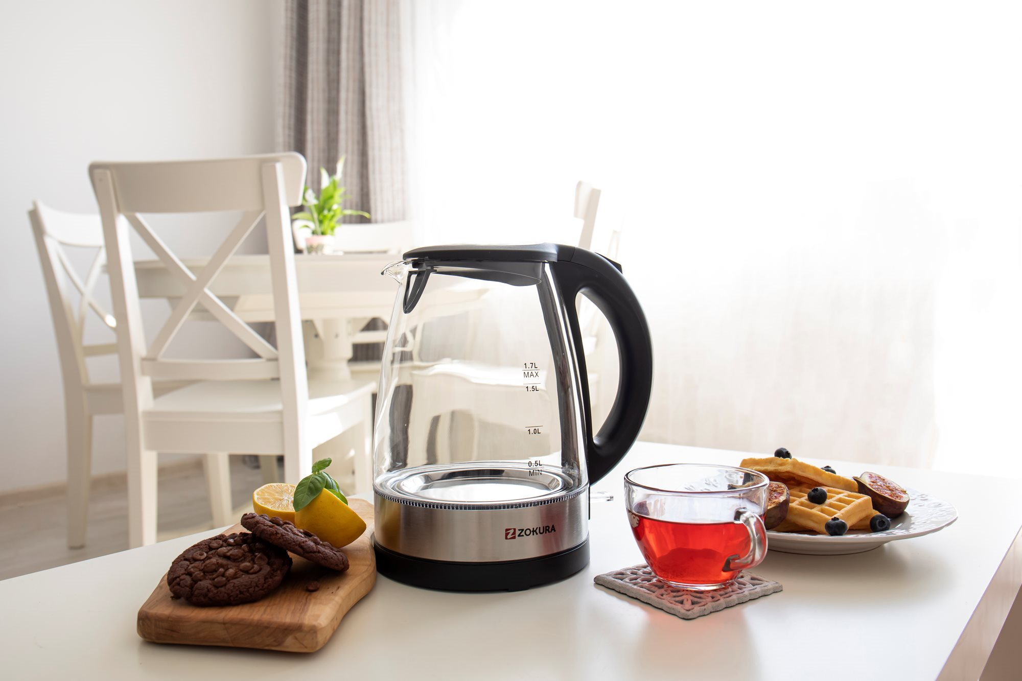 Egy asztal vízforralóval, egy csésze teával, kekszekkel és gofrikkal. Háttérben egy ablak és fehér székek.