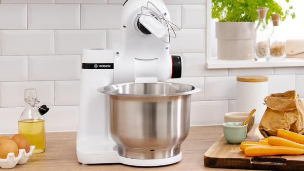 Weißer Küchenmixer mit Schüssel und Schneebesen, Lebensmittel auf der Theke.
