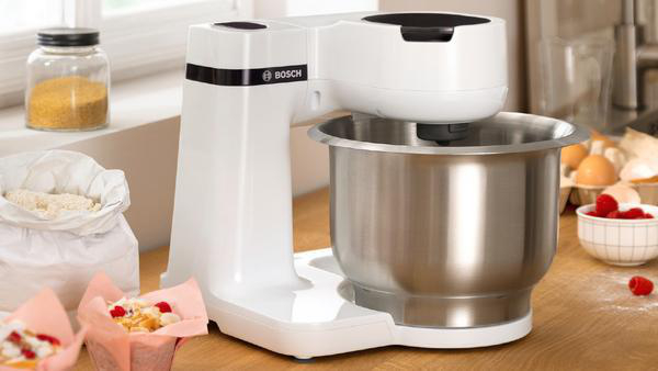 Weißer Bosch-Küchenmixer mit Edelstahlschüssel, Mehl und Muffins auf dem Tisch.
