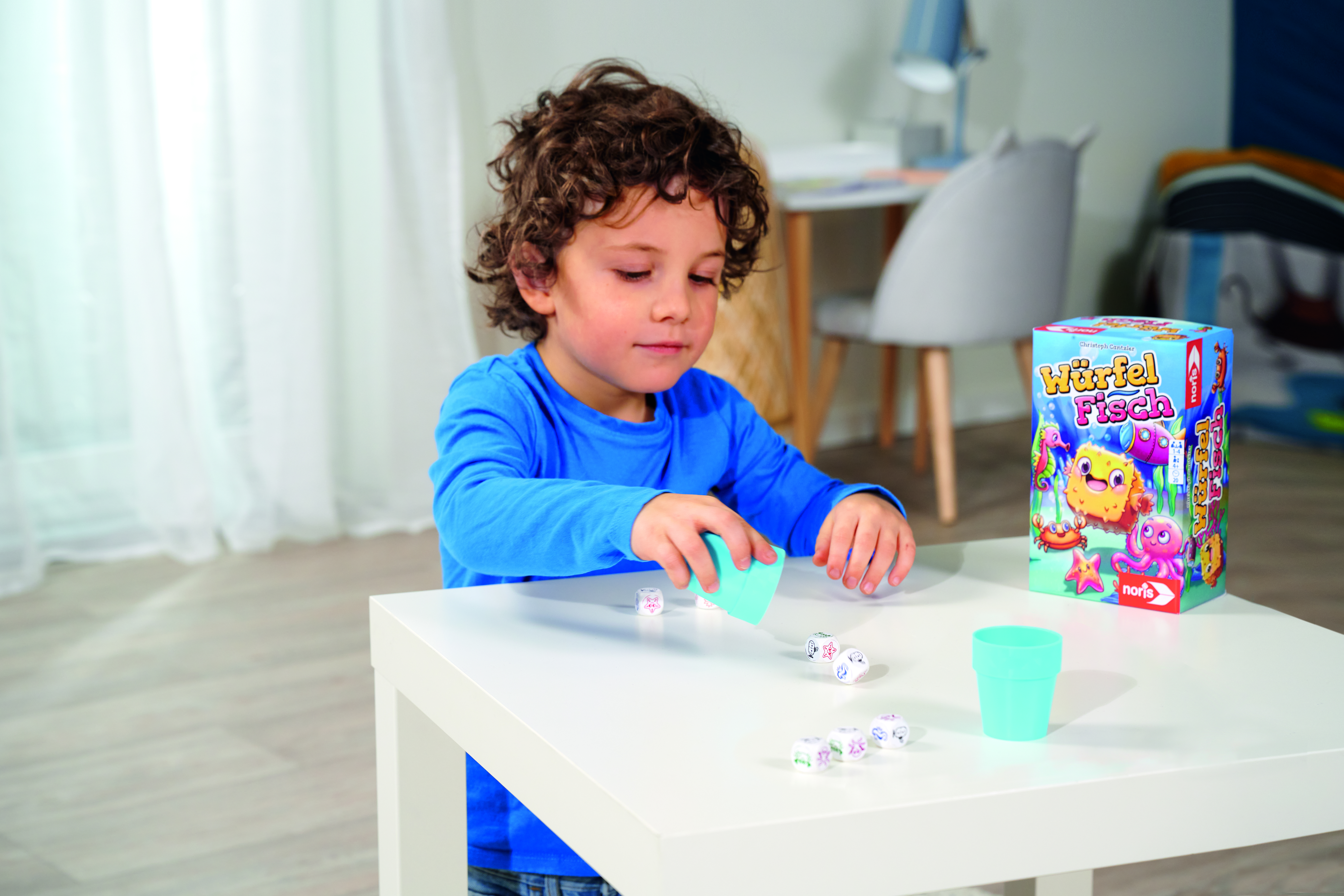 Ein Junge spielt ein Würfelspiel mit Würfeln und einem Becher, die Spielbox ist sichtbar.
