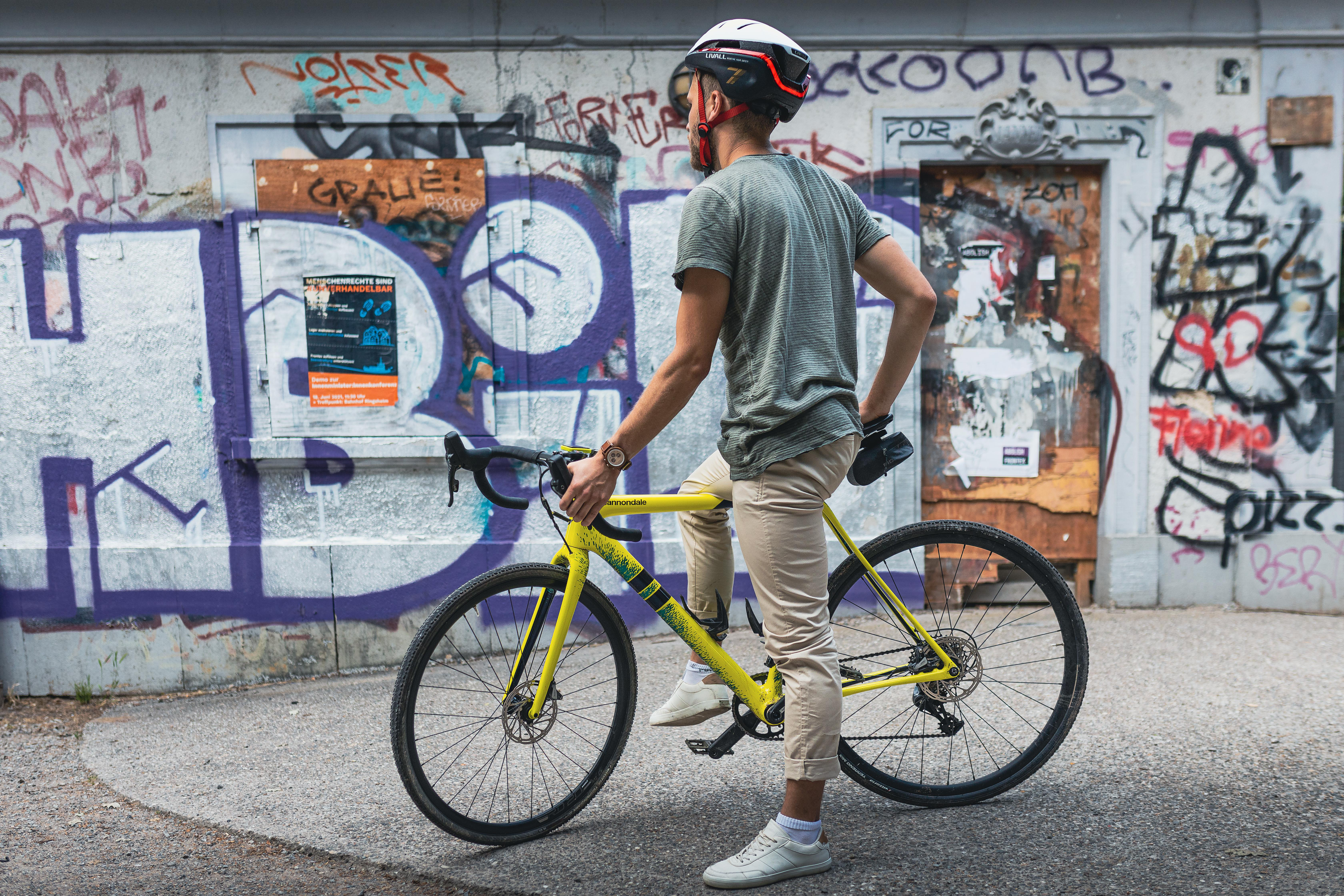 Ein Mann mit Helm steht neben einem gelben Fahrrad vor einer Graffiti-Wand.