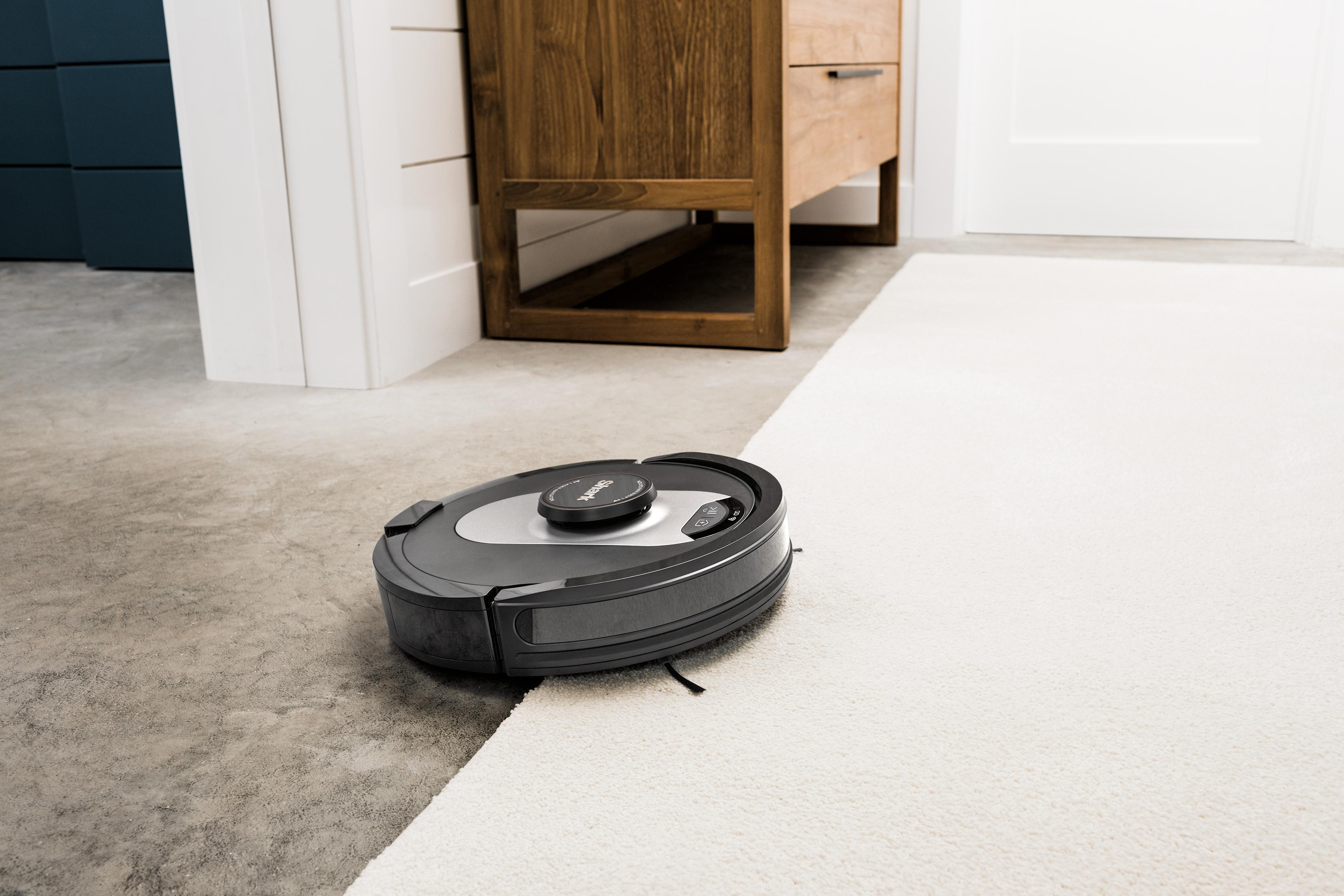 Ein Roboter-Staubsauger reinigt einen Boden mit einem Übergang zwischen Teppich und grauen Fliesen.