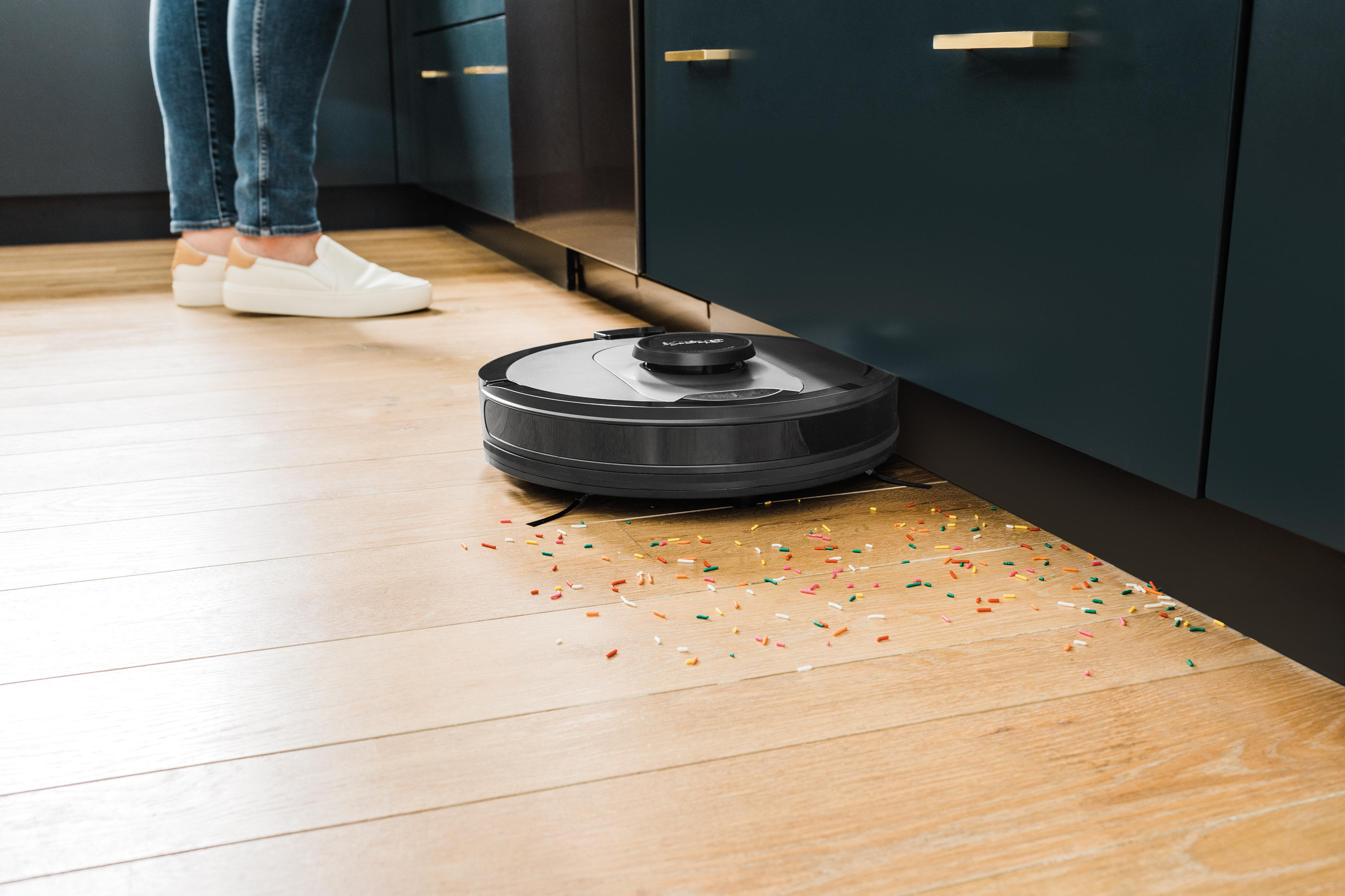 Roboter-Staubsauger auf Holzboden mit Konfetti, in der Nähe eines Schranks und Beinen einer Person.