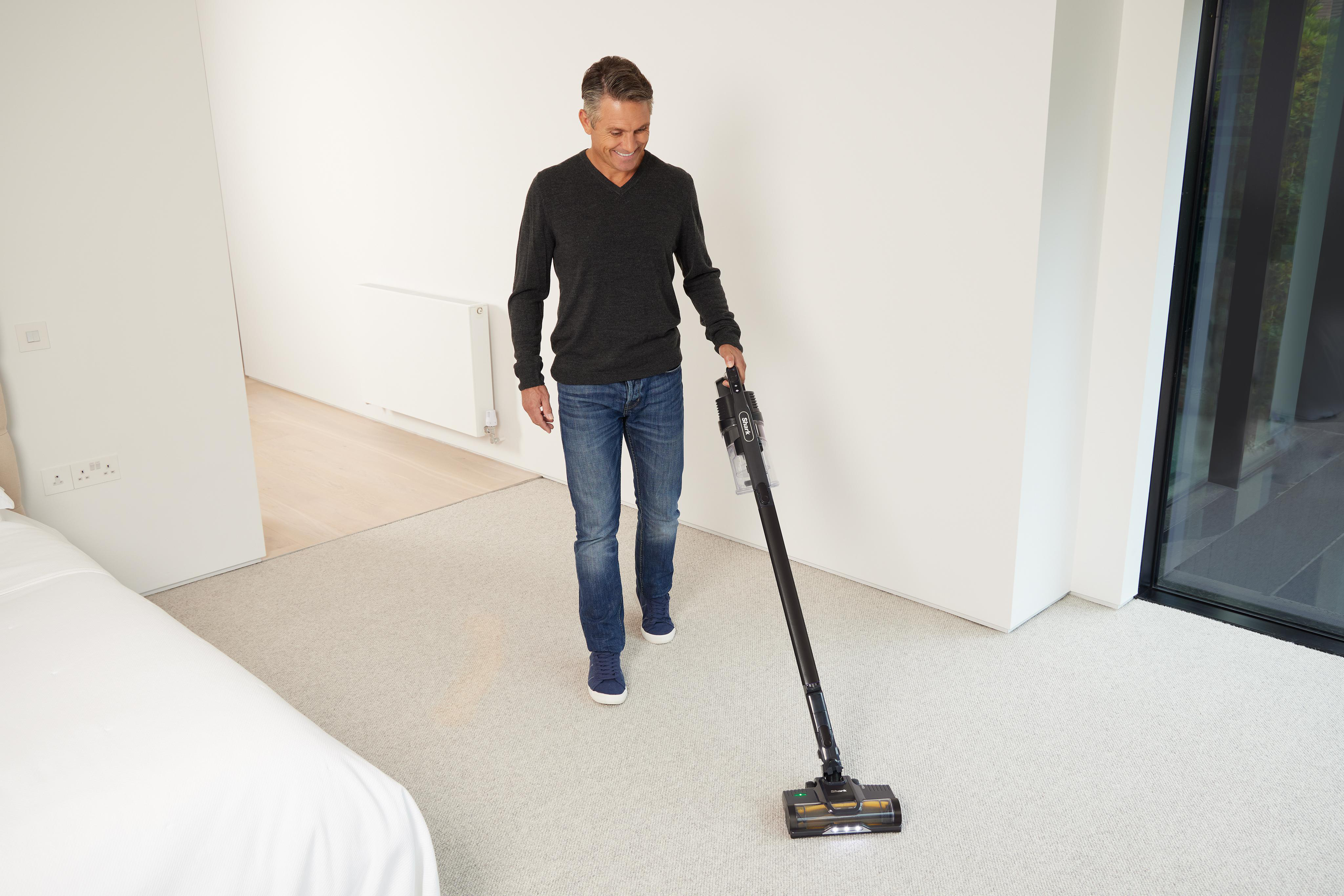 Ein Mann, der ein Zimmer mit weißem Teppich saugt und einen kabellosen Staubsauger hält.