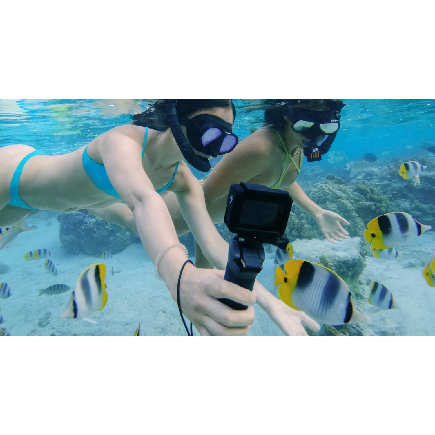 Dos personas bucean bajo el agua con una cámara, rodeadas de peces y coral. Agua azul brillante y sol.