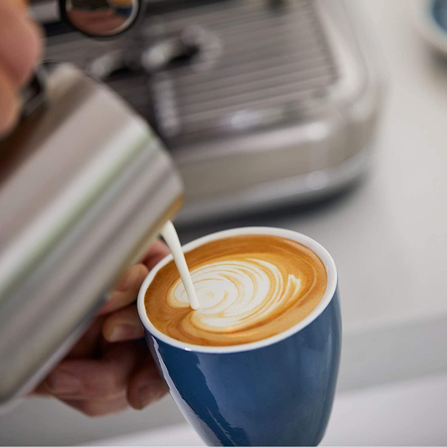 Una persona vierte leche en una taza de café azul, creando arte latte.