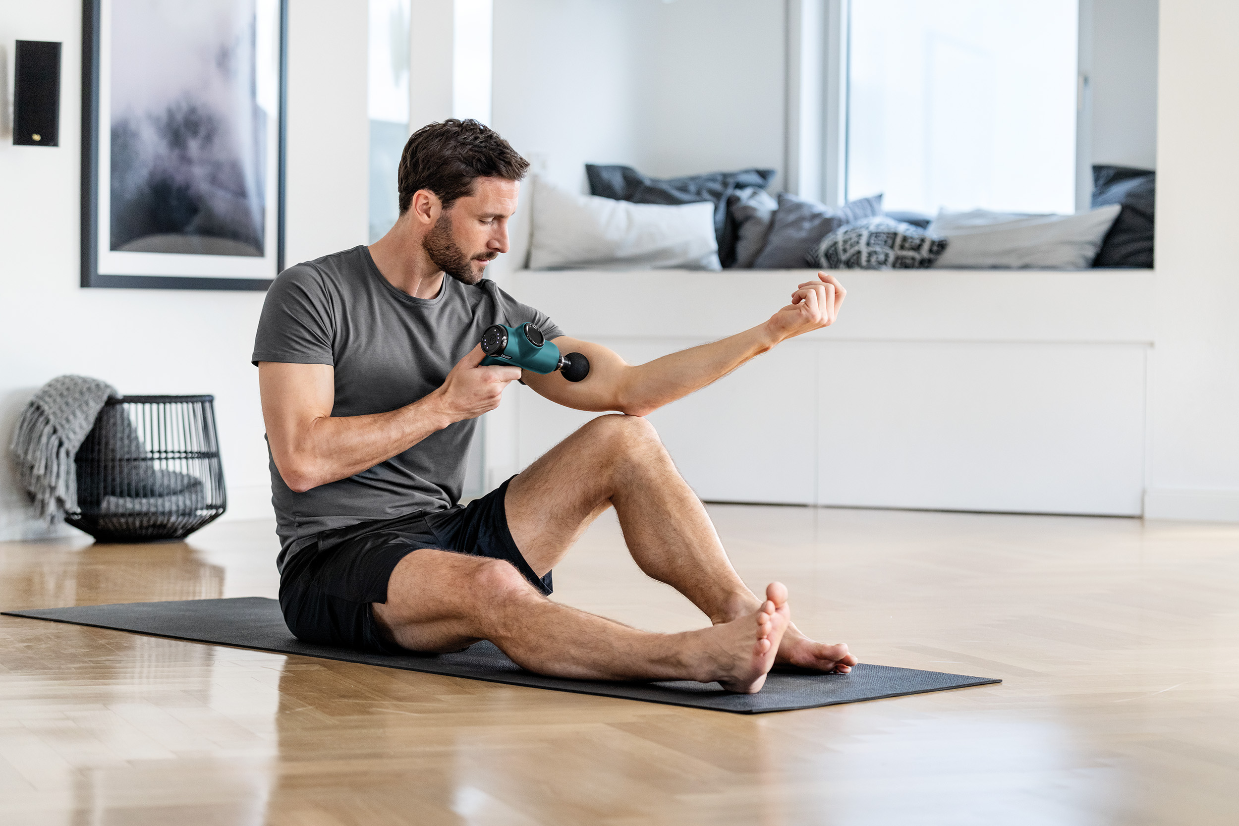 Ein Mann sitzt und benutzt eine Massagepistole an seinem Arm. Er ist auf einer Matte. Der Hintergrund ist der Raum.