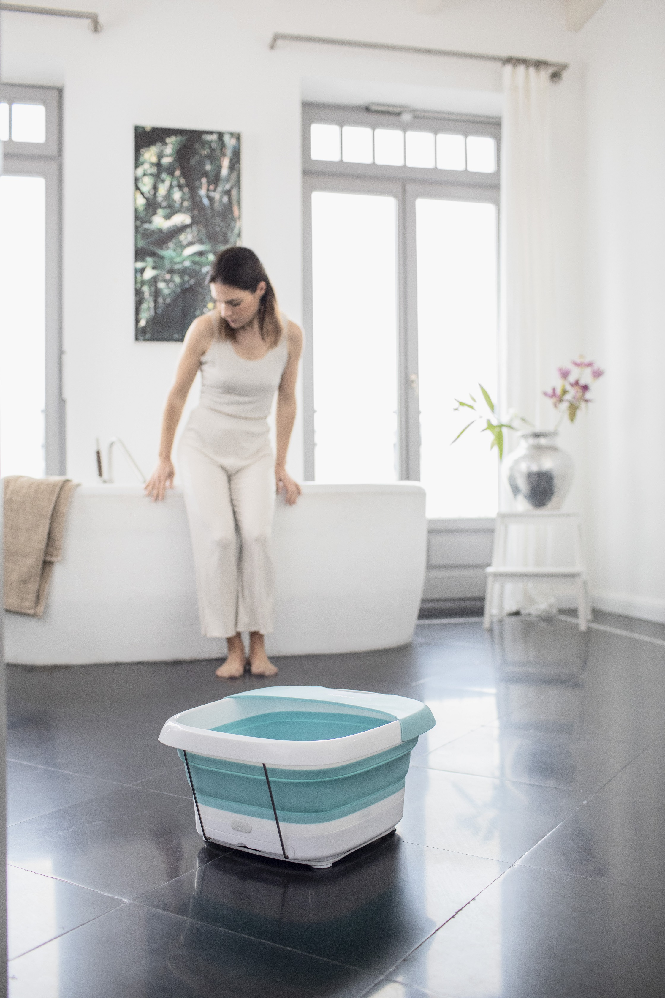 Woman in a bathroom, near a bathtub. A foot spa is on the floor in the foreground.