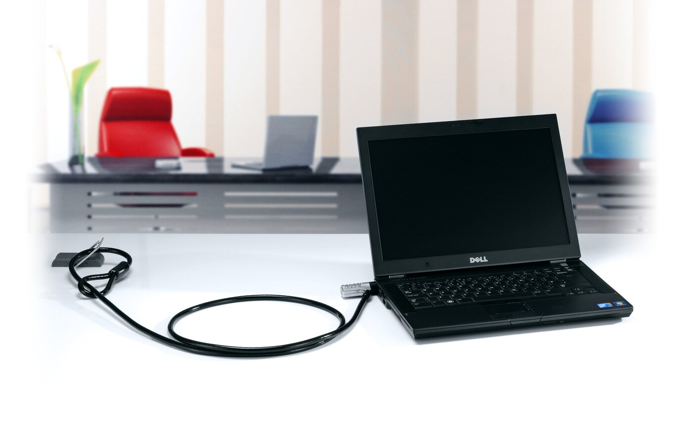 Egy laptop kábellel és zárral rögzítve egy fehér asztalon. Van egy monitor és egy másik laptop.