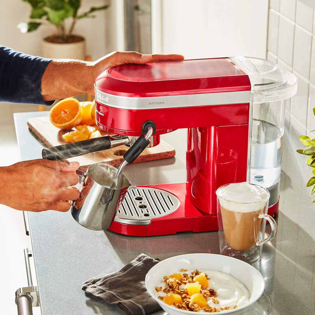 Egy piros KitchenAid eszpresszó gép, amint egy személy tejet önt egy csészébe, egy joghurtos tál mellett.