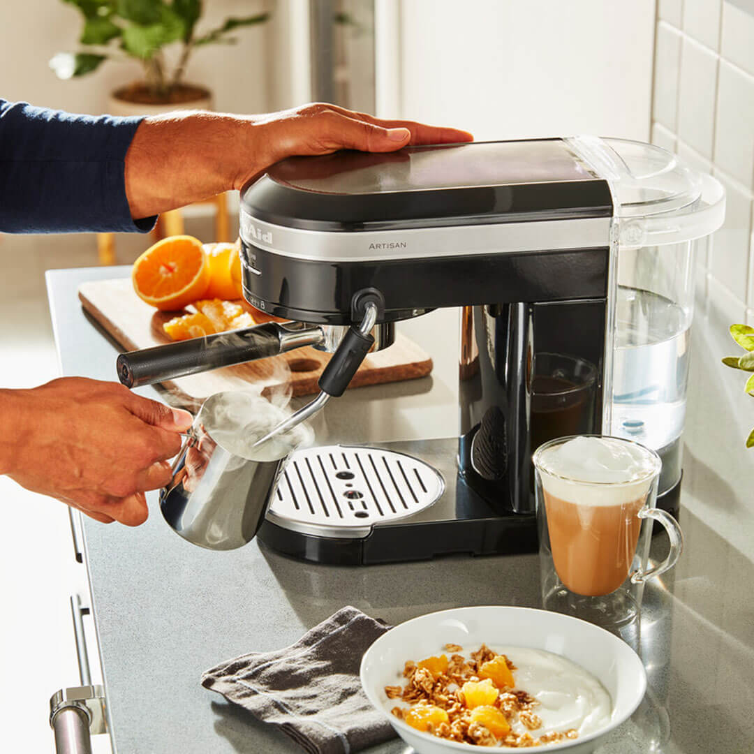 Egy személy KitchenAid eszpresszó gépet használ kávéital készítéséhez, reggeli elemekkel.