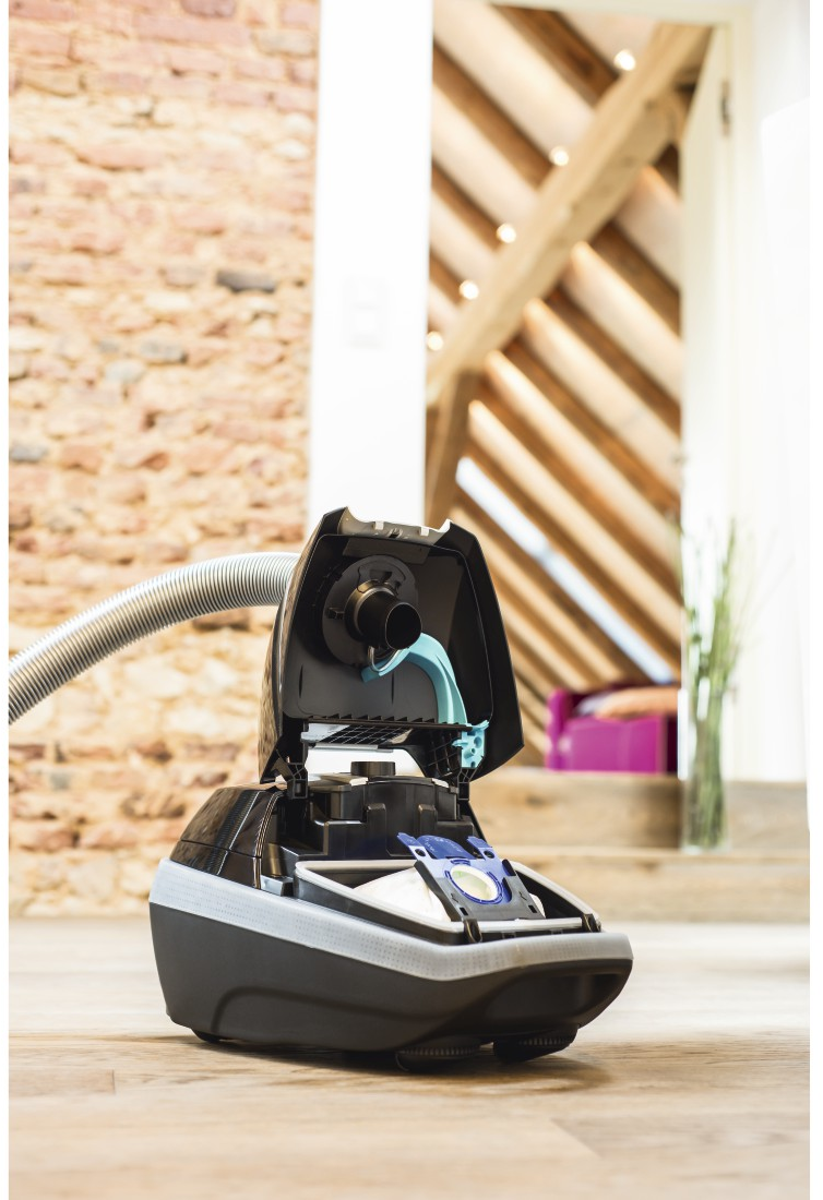 Un aspirateur ouvert, montrant le sac et le filtre. Sol en bois et mur de briques en arrière-plan.