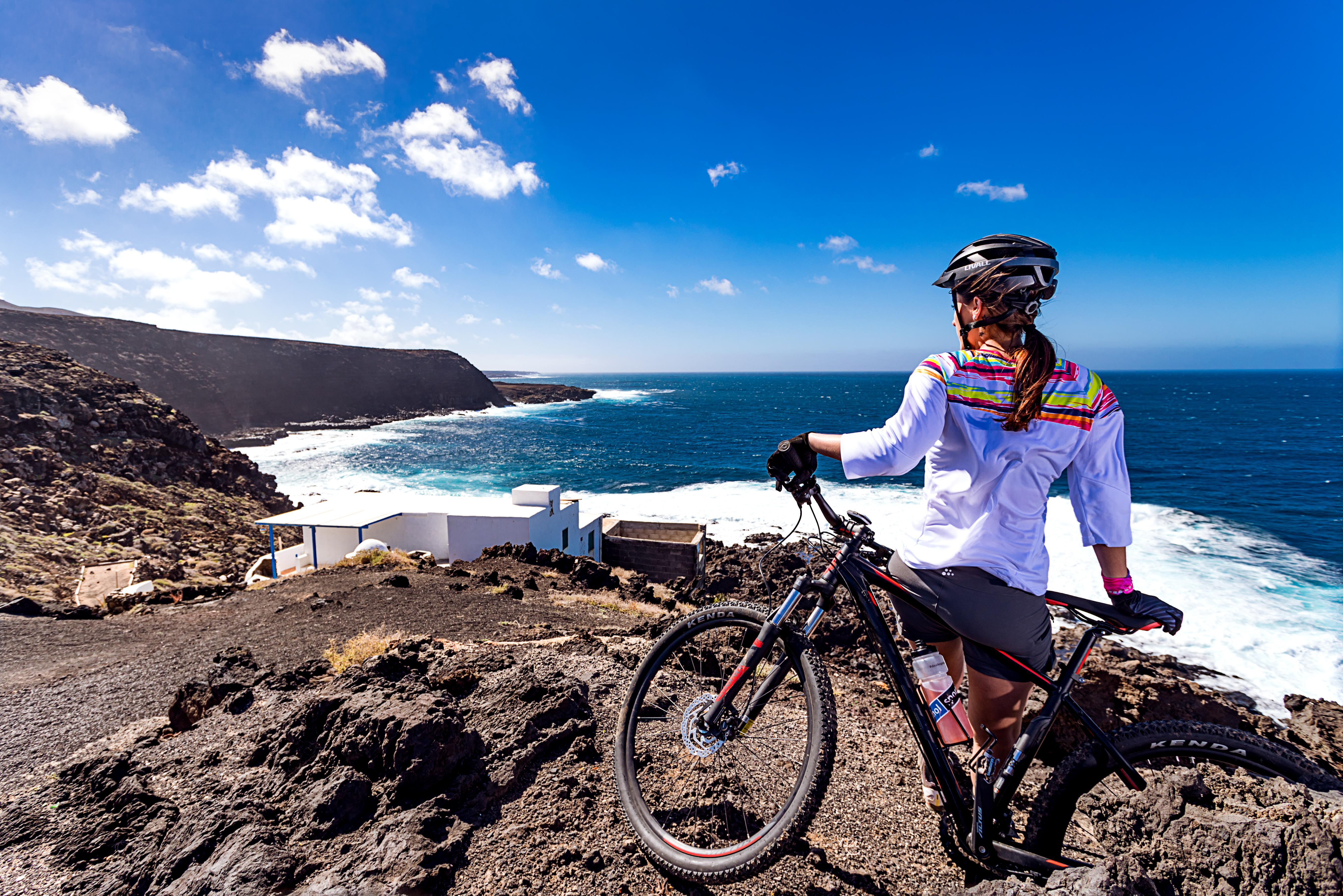 Frau auf Mountainbike blickt aufs Meer, blauer Himmel, sonniger Tag, Küstenansicht.