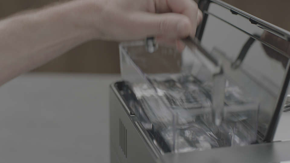 A hand opening a clear plastic container inside a coffee machine, holding packets.