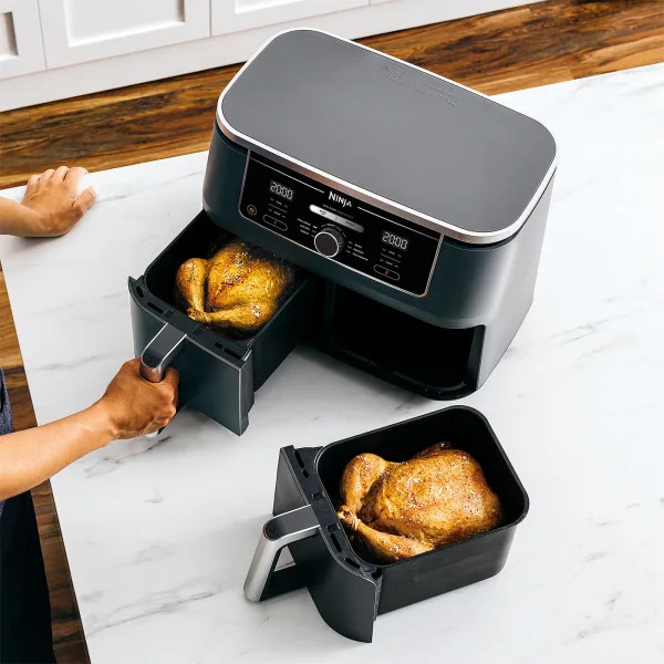 Une friteuse à air avec deux paniers, chacun avec un poulet cuit. Une personne tient un panier.
