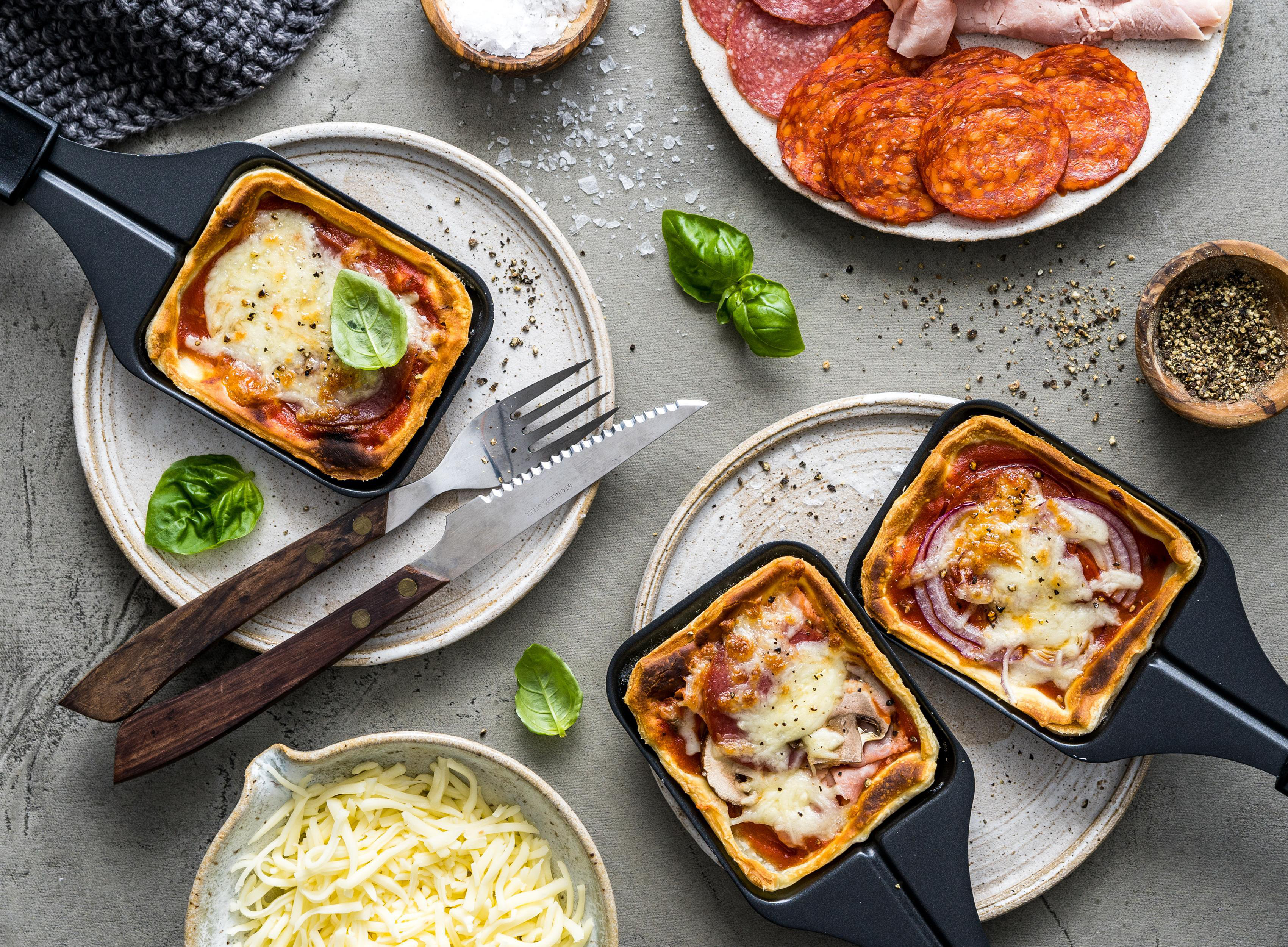 Drei Mini-Pizzas in schwarzen Pfannen mit Belägen, umgeben von Zutaten.