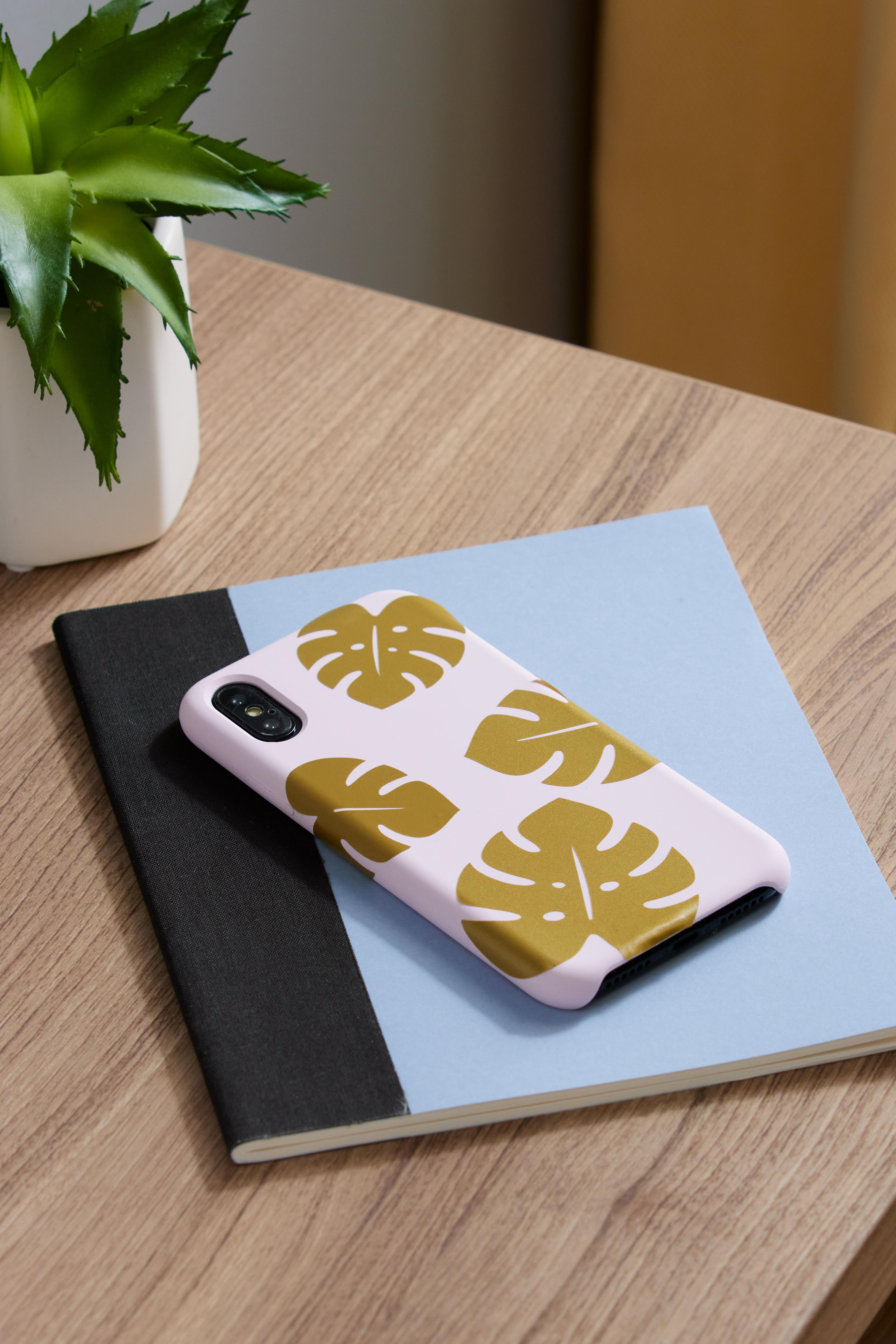 Eine Handyhülle mit Monstera-Blattmuster auf einem Notizblock und einem Tisch.