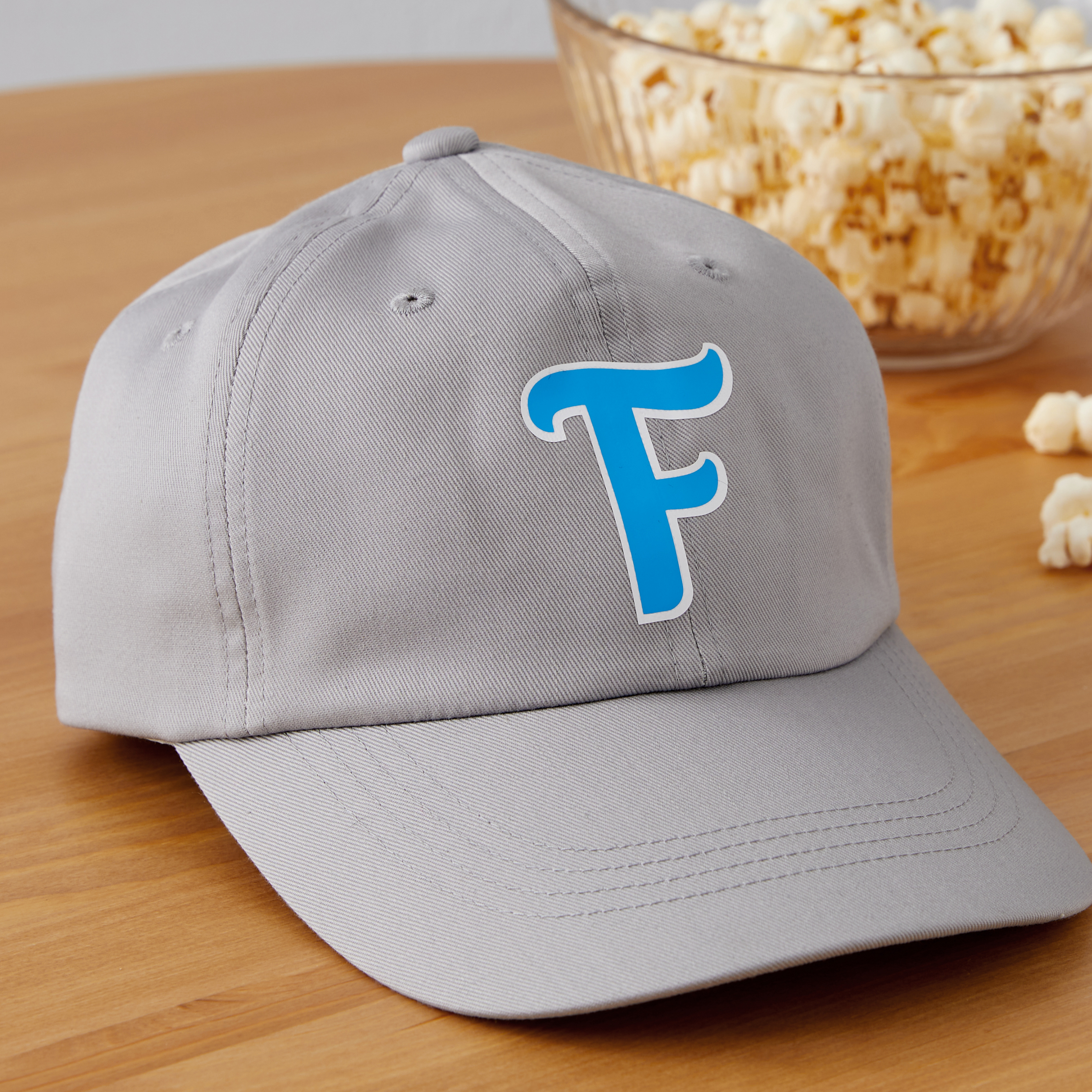 Eine graue Baseballkappe mit blauem 'F'-Logo liegt auf einem Holztisch. Im Hintergrund eine Schüssel Popcorn.