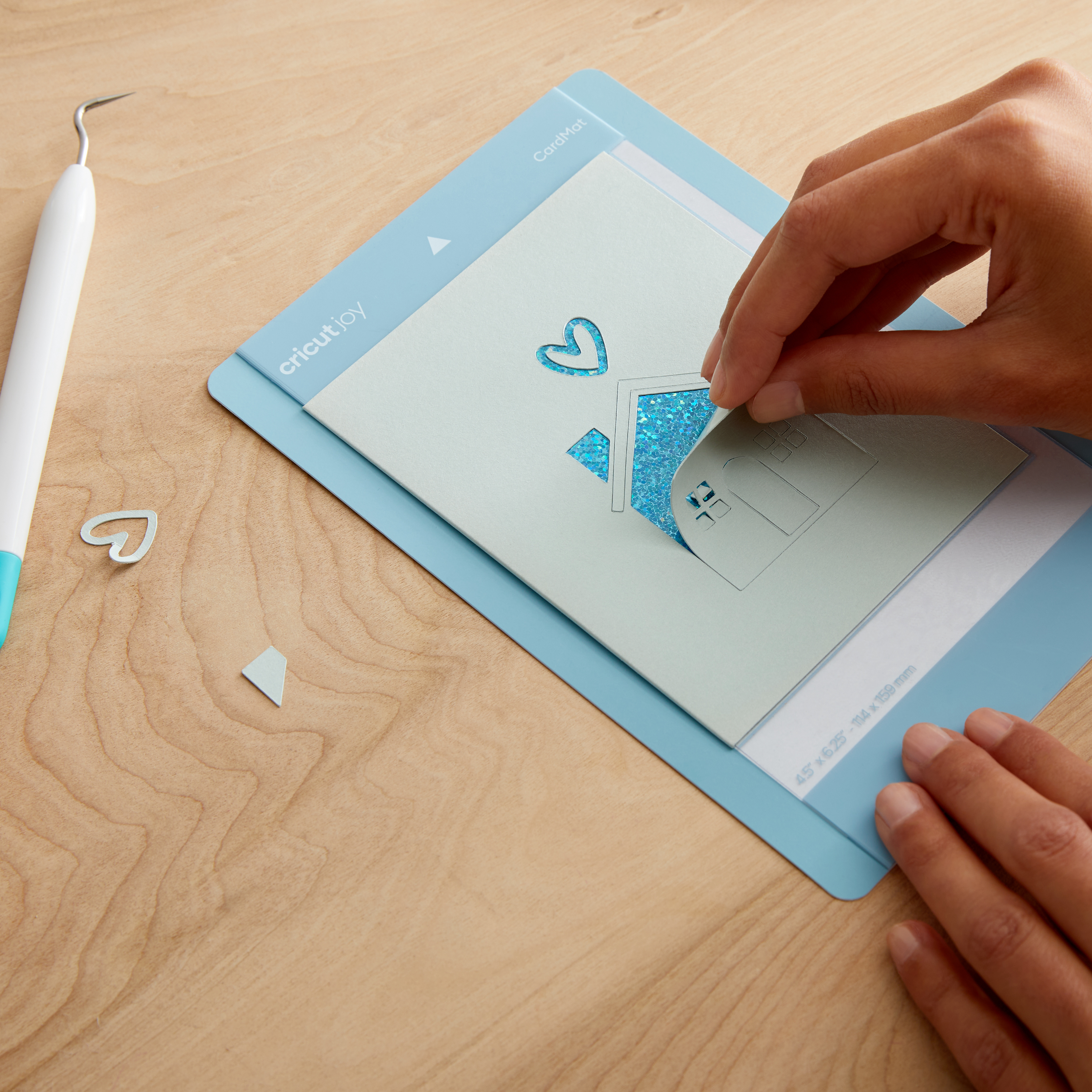 Hände, die ein Haus aus Papier von einer blauen Schneidematte abziehen. Ein Bastelmesser und ein Herz-Ausschnitt liegen daneben.