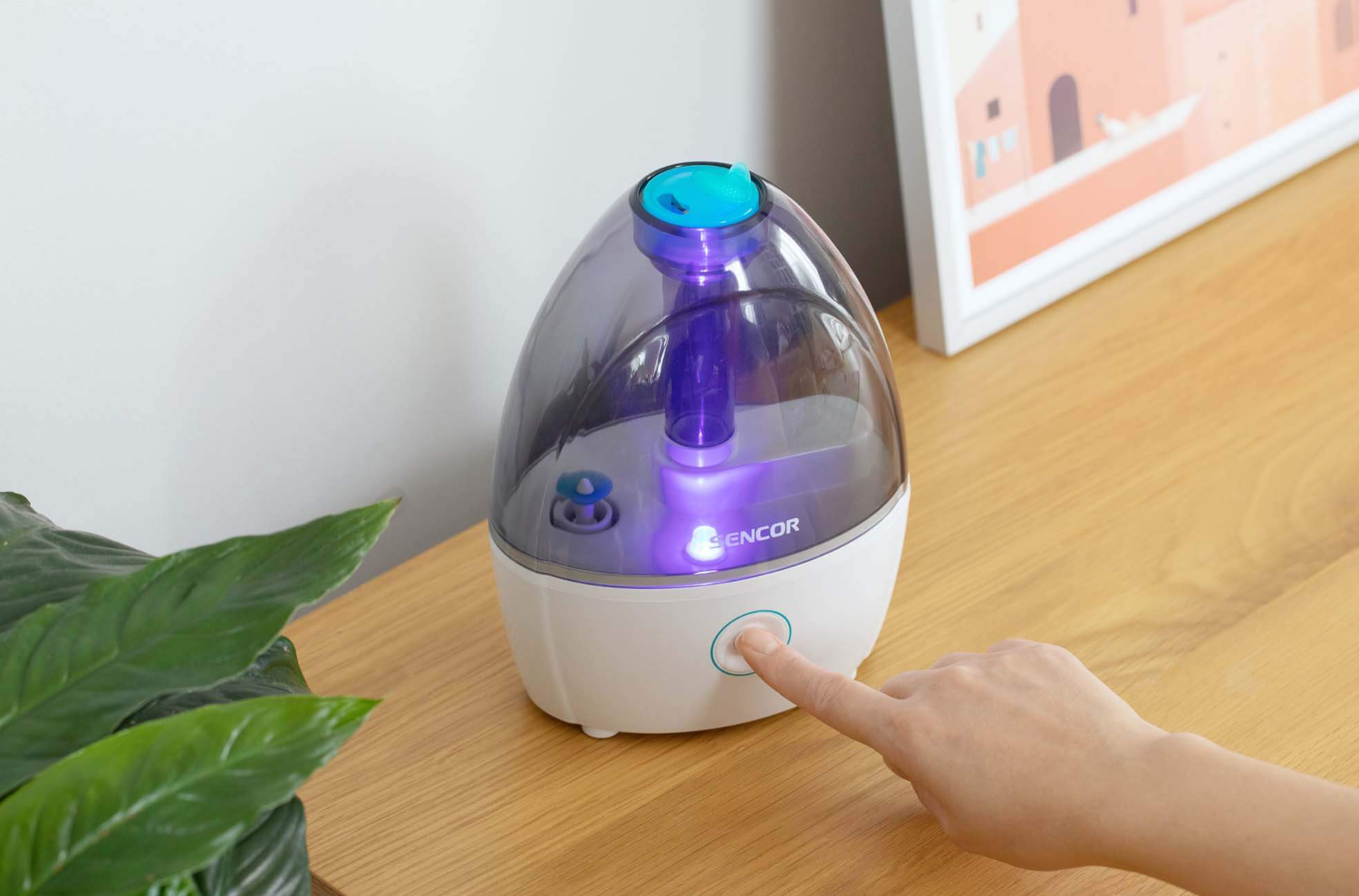 Párásító fehér alappal, átlátszó testtel és kék elemekkel, kéz a gombot érinti.
